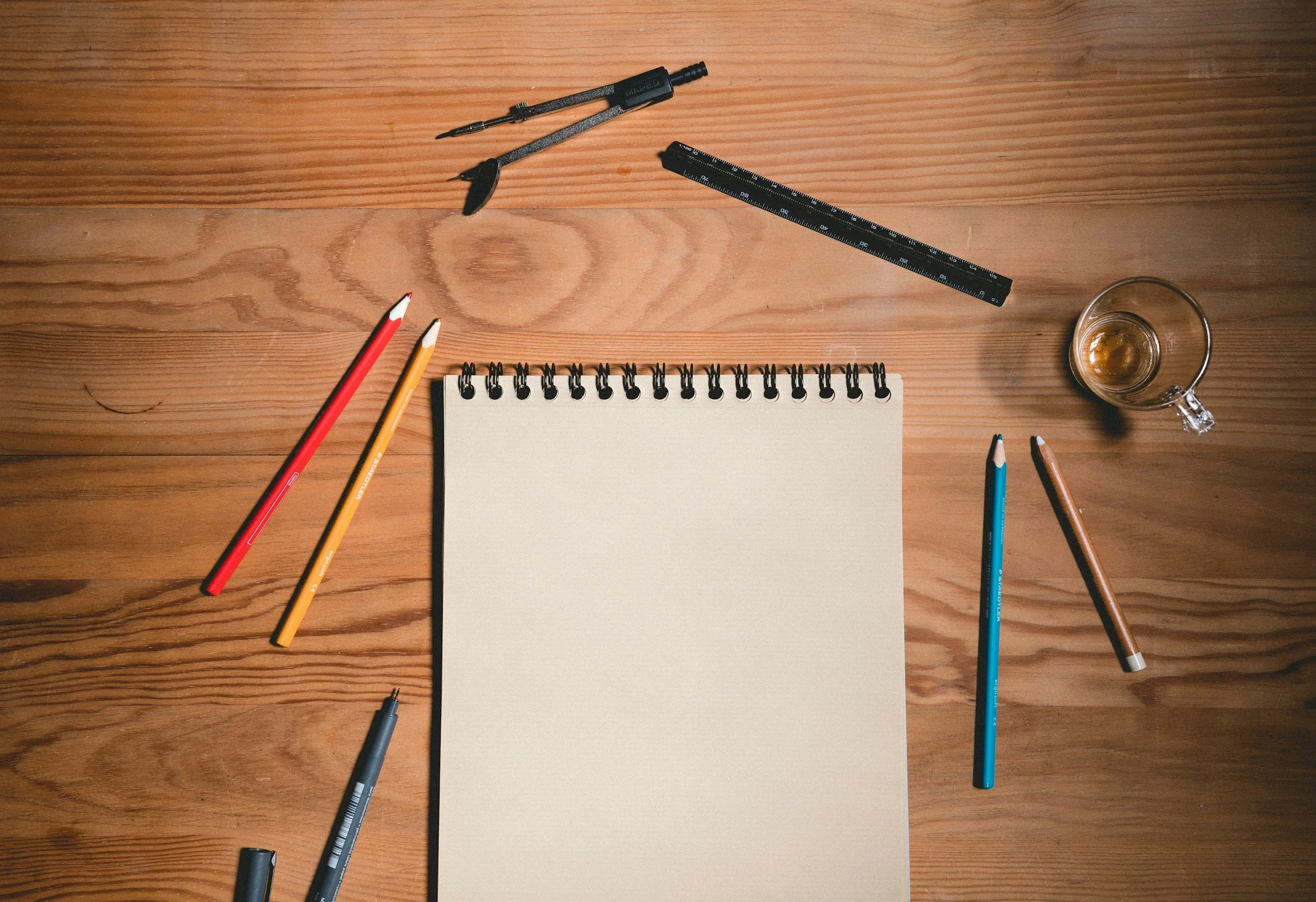 A wooden desk has a spiral notepad, colorful pens, a glass cup with a beverage, a ruler, a screwdriver set, and several other pens and markers scattered across it. This denotes a sketchbook before anything is drawn on