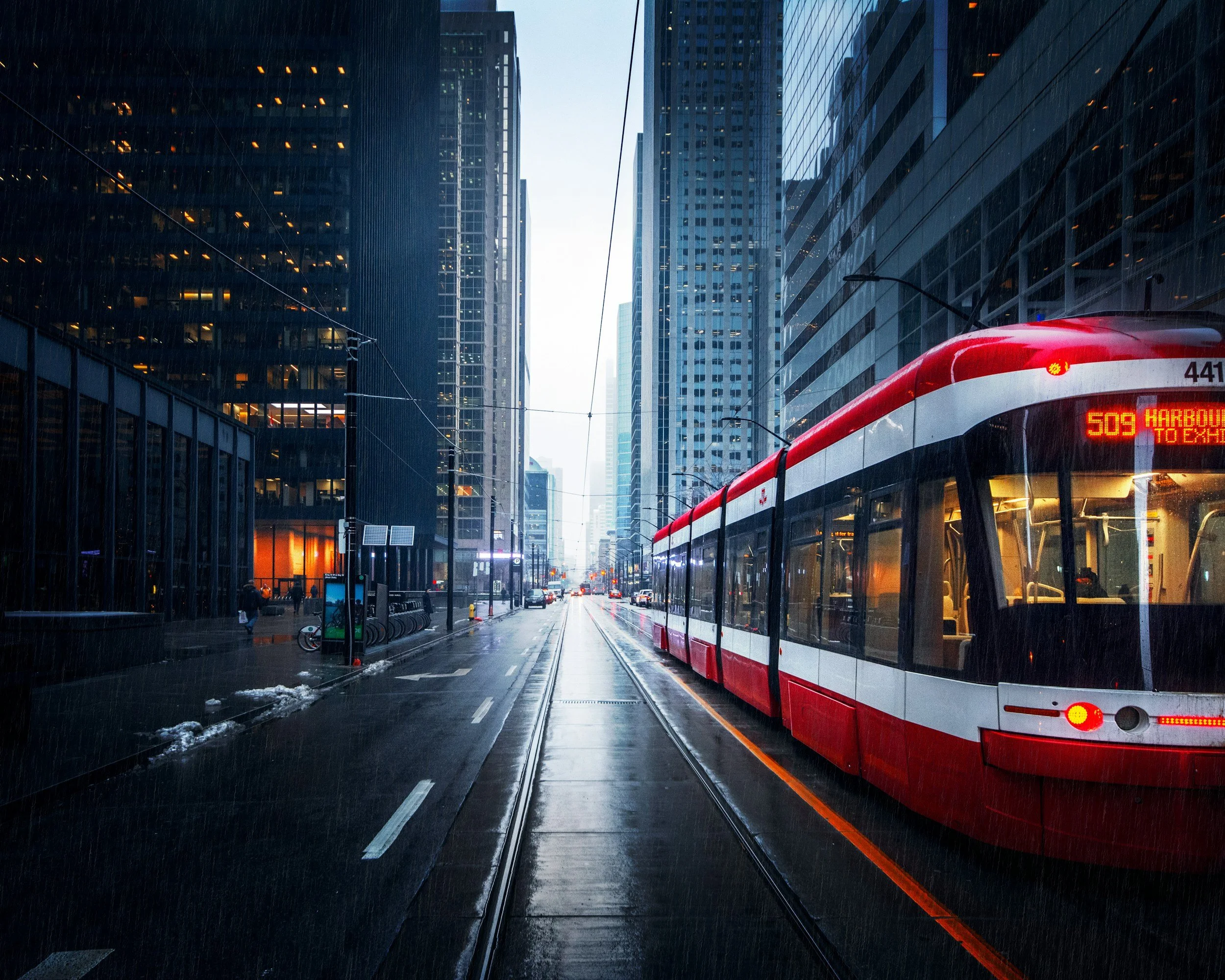 A Toronto city street with tall glass skyscrapers on both sides under rainy weather. A red and white tram is moving along the tracks, with a digital sign displaying the route number 509 and destinations Harbor to T.