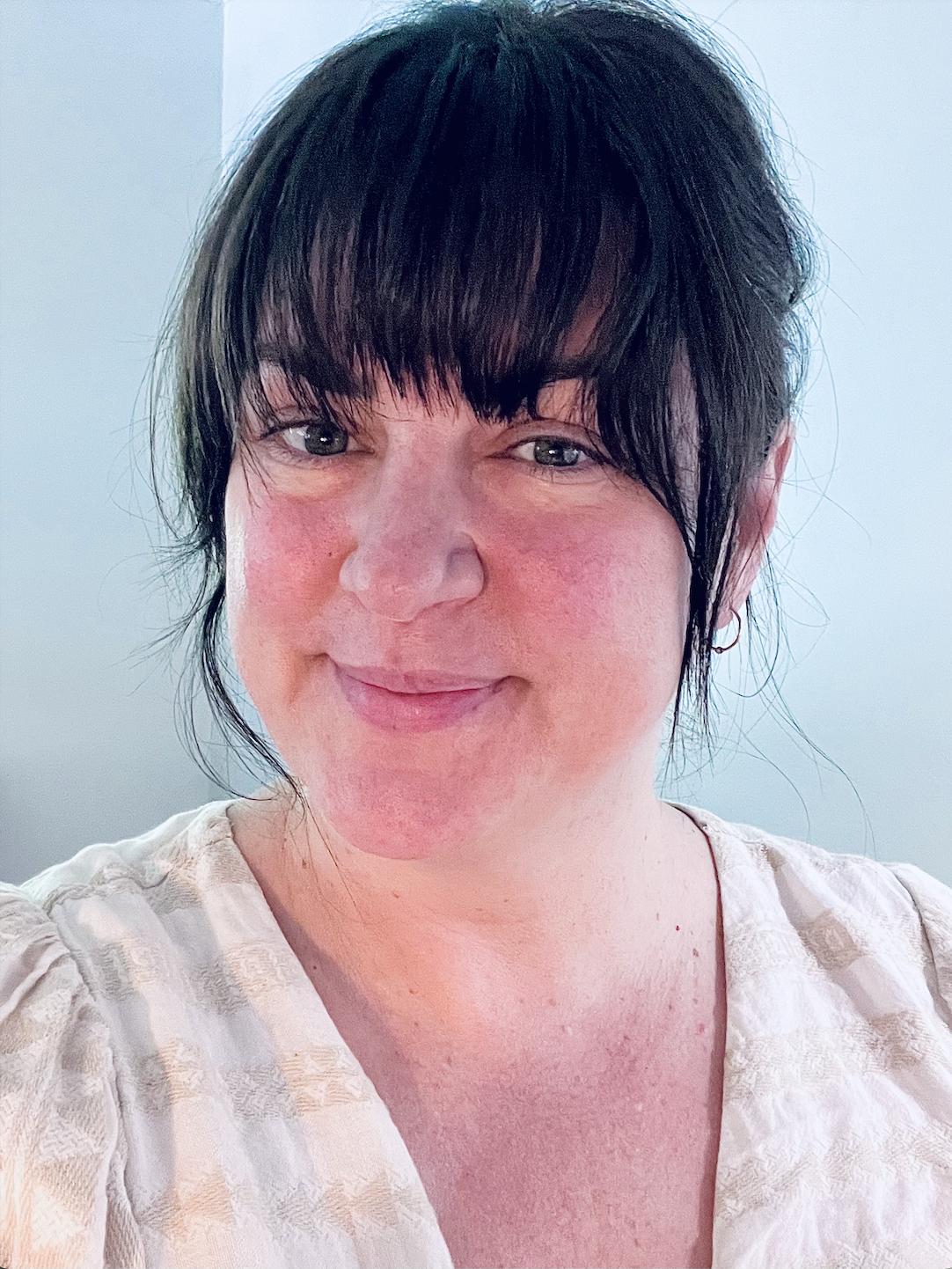 Close-up of Vicki Sloot with tied back brown hair, pale skin, green eyes, and a slight smile, wearing a light-colored patterned blouse against a light background.
