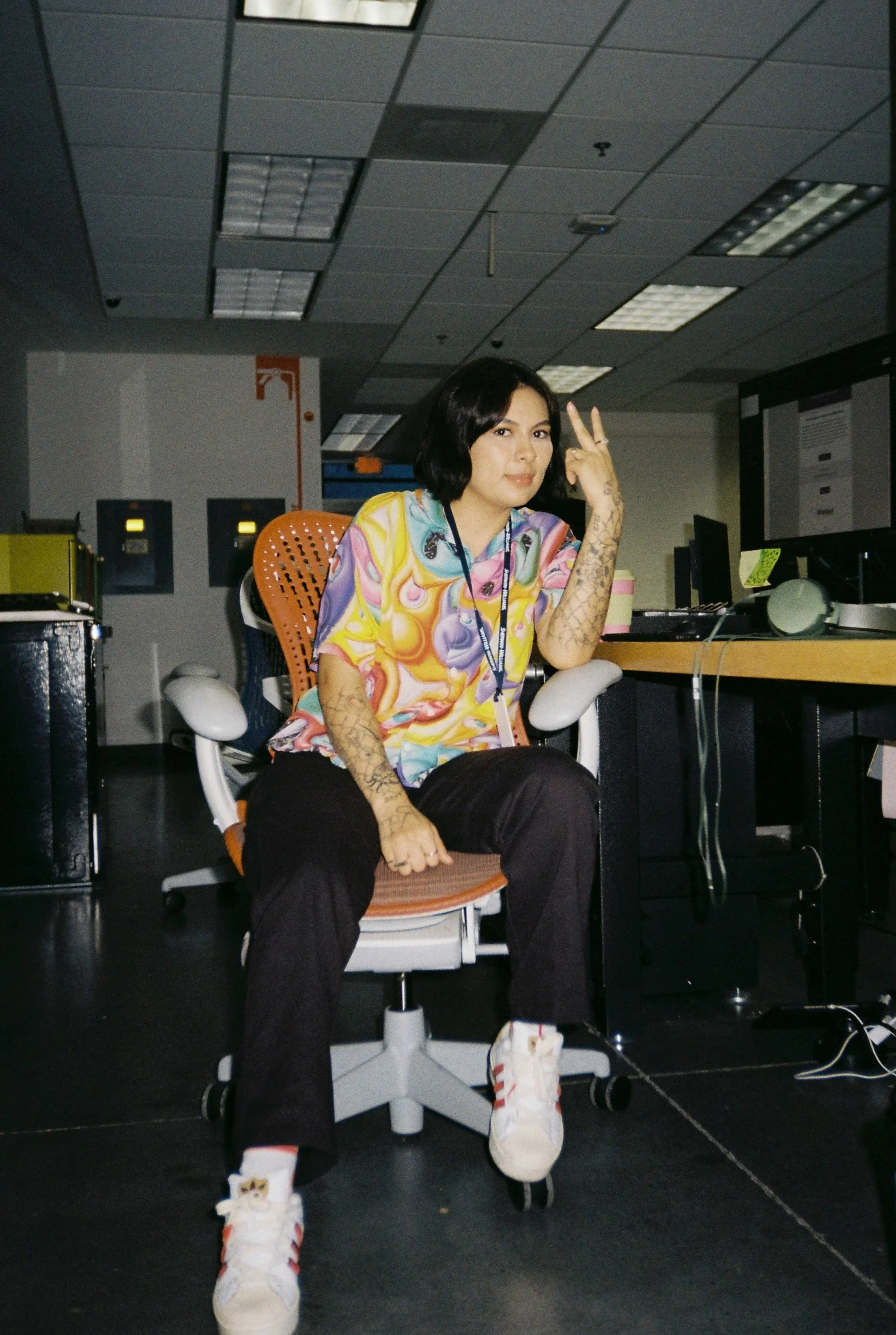 A woman with short dark hair and tattoos on her arms, wearing a colorful patterned shirt and white sneakers with cat face designs, sitting on an office chair in an office environment, making a peace sign with her right hand.