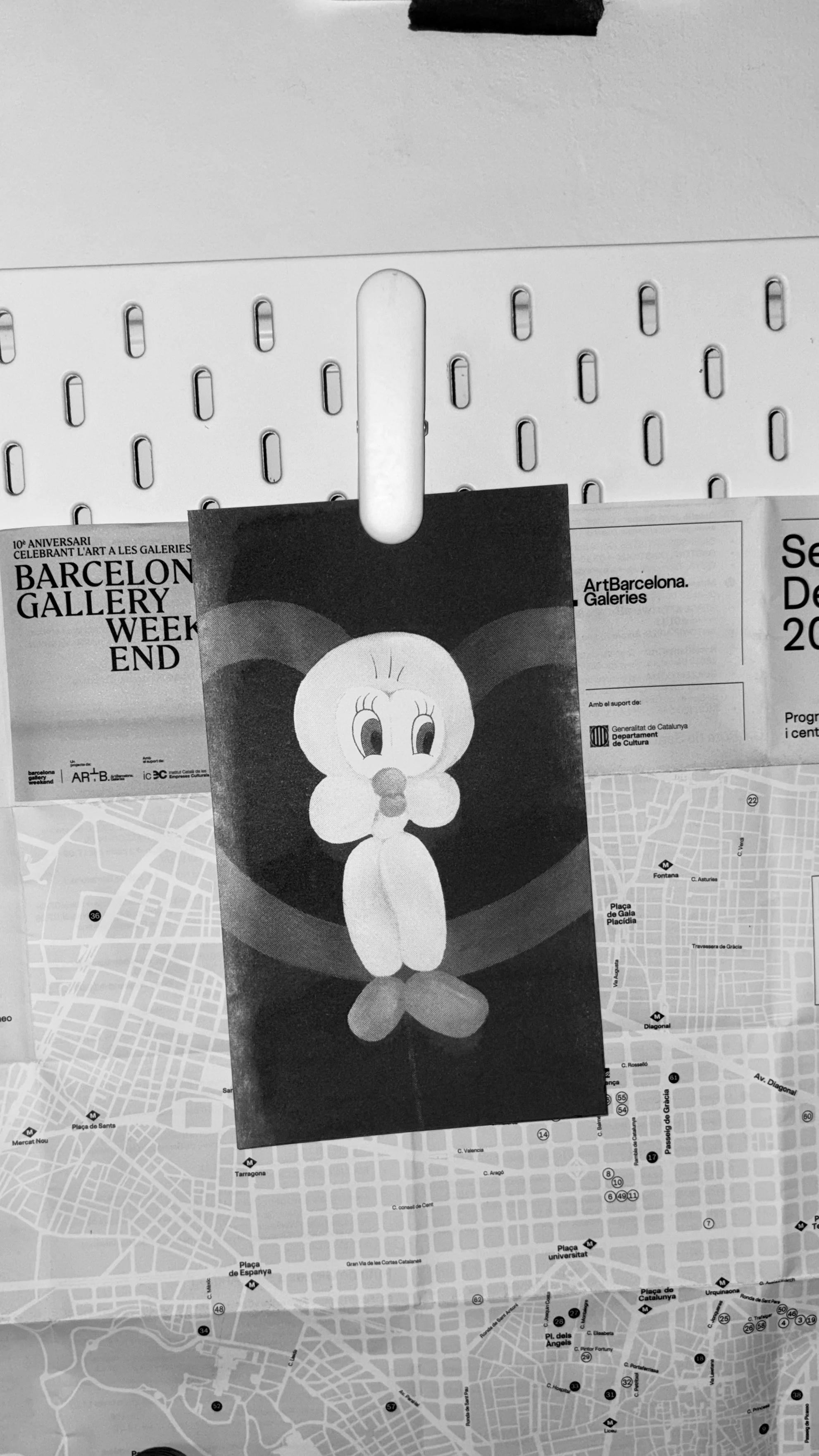 Black and white photo of a bulletin board with attached papers, including a map of Barcelona with street names, a flyer for Barcelona Gallery Weekend, and a cartoon image of a dog with large eyes, a small nose, and big ears.