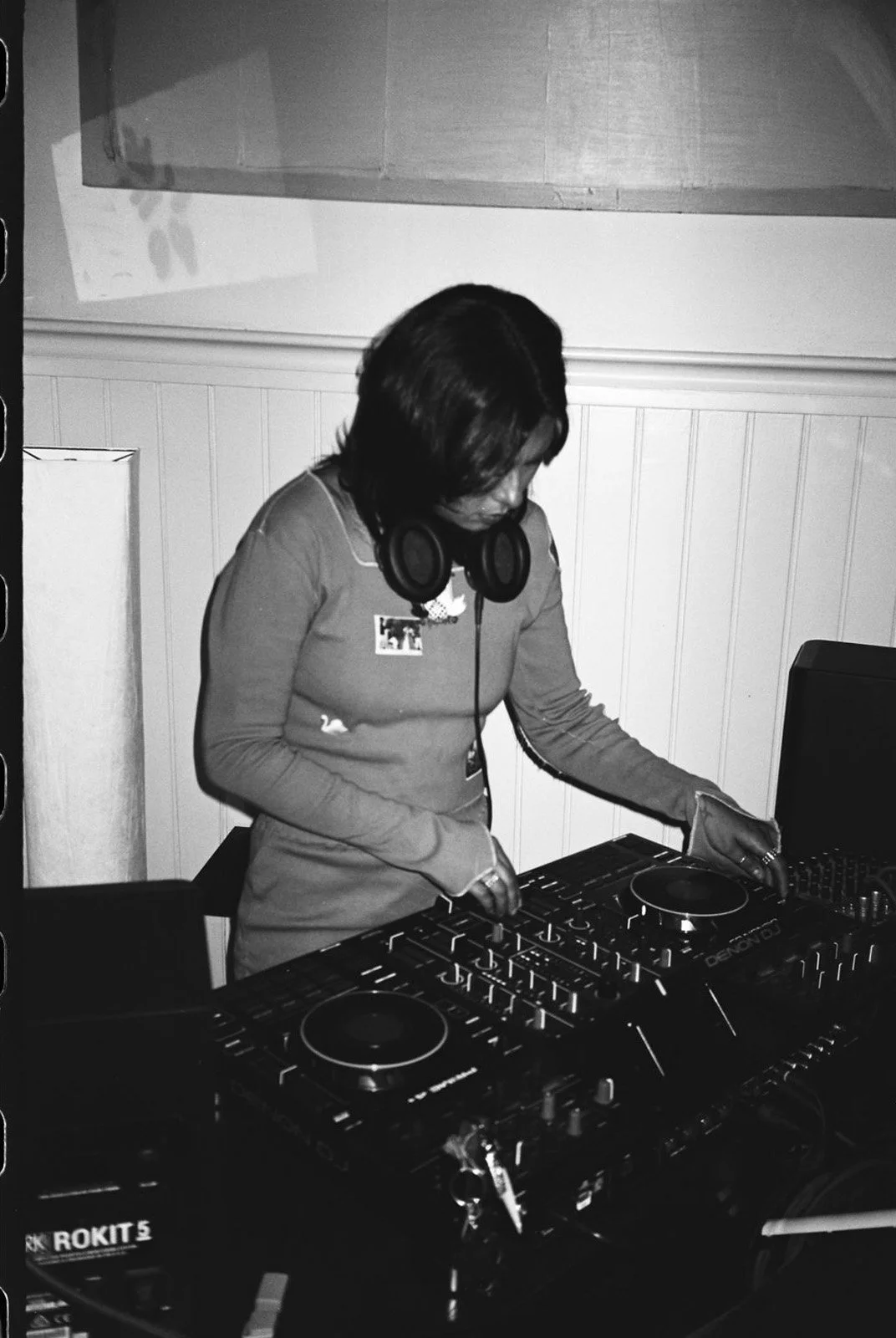 A woman with headphones around her neck DJing with a mixer and turntables in a dimly lit room.