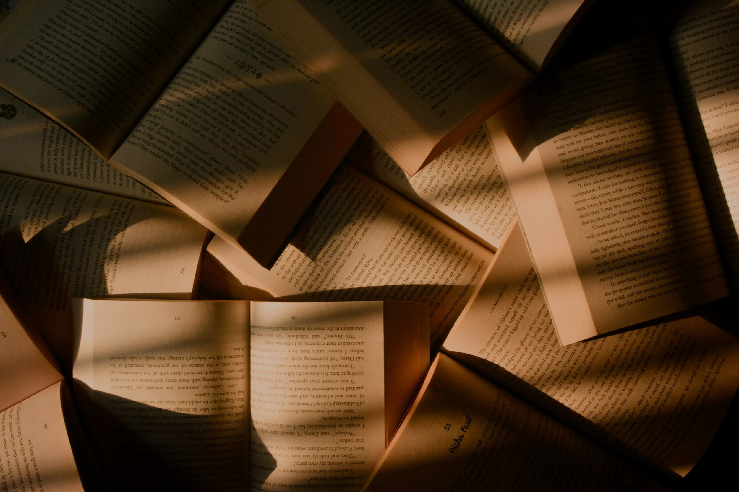 Multiple open books arranged on a surface, illuminated with warm light, with visible pages of text.