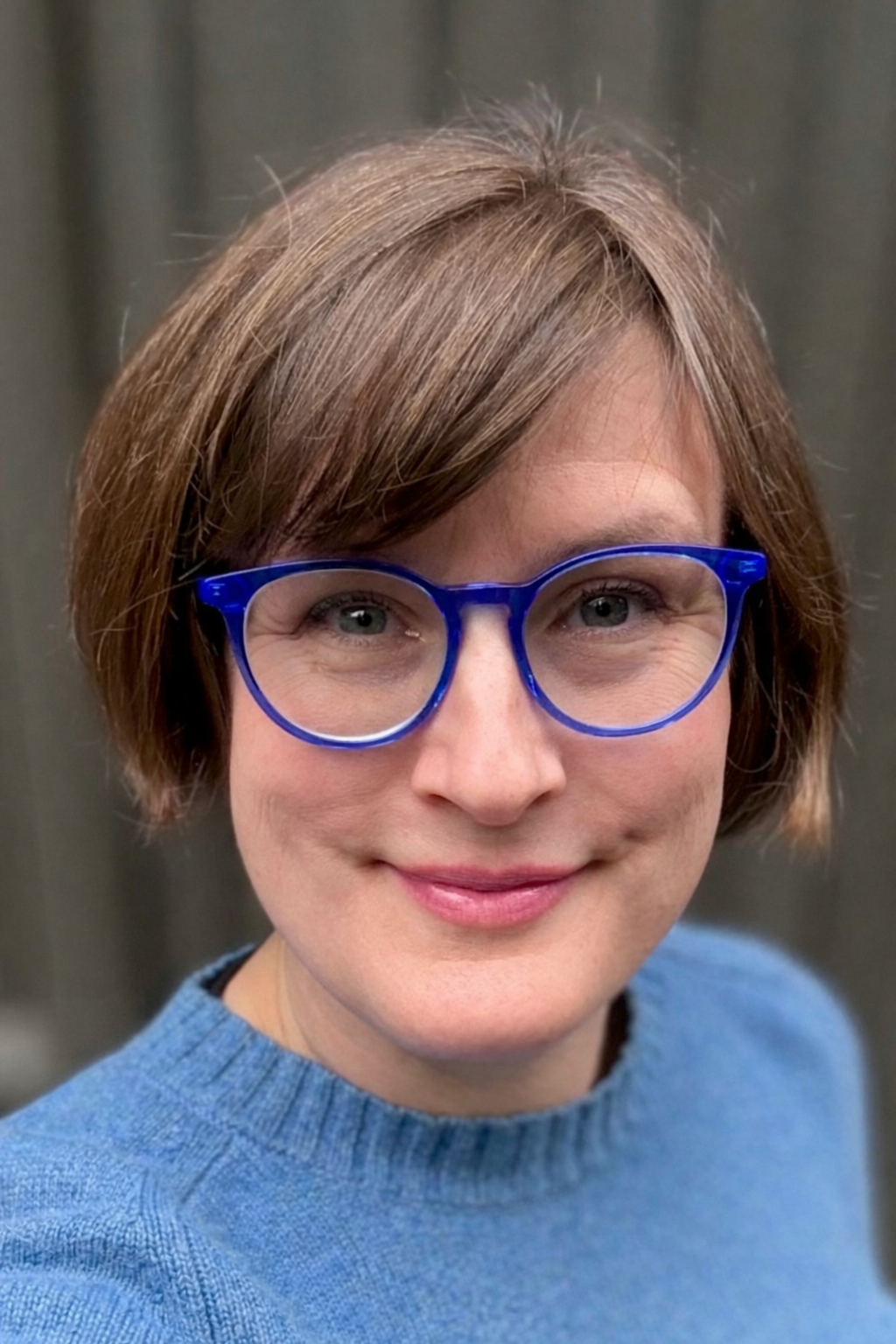 A woman with short brown hair wearing blue glasses and a blue sweater, smiling at the camera.