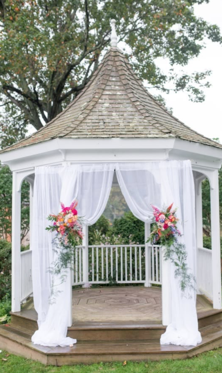Decorated white gazebo with sheer white curtains and vibrant flower arrangements at the entrance, set in a garden with trees.