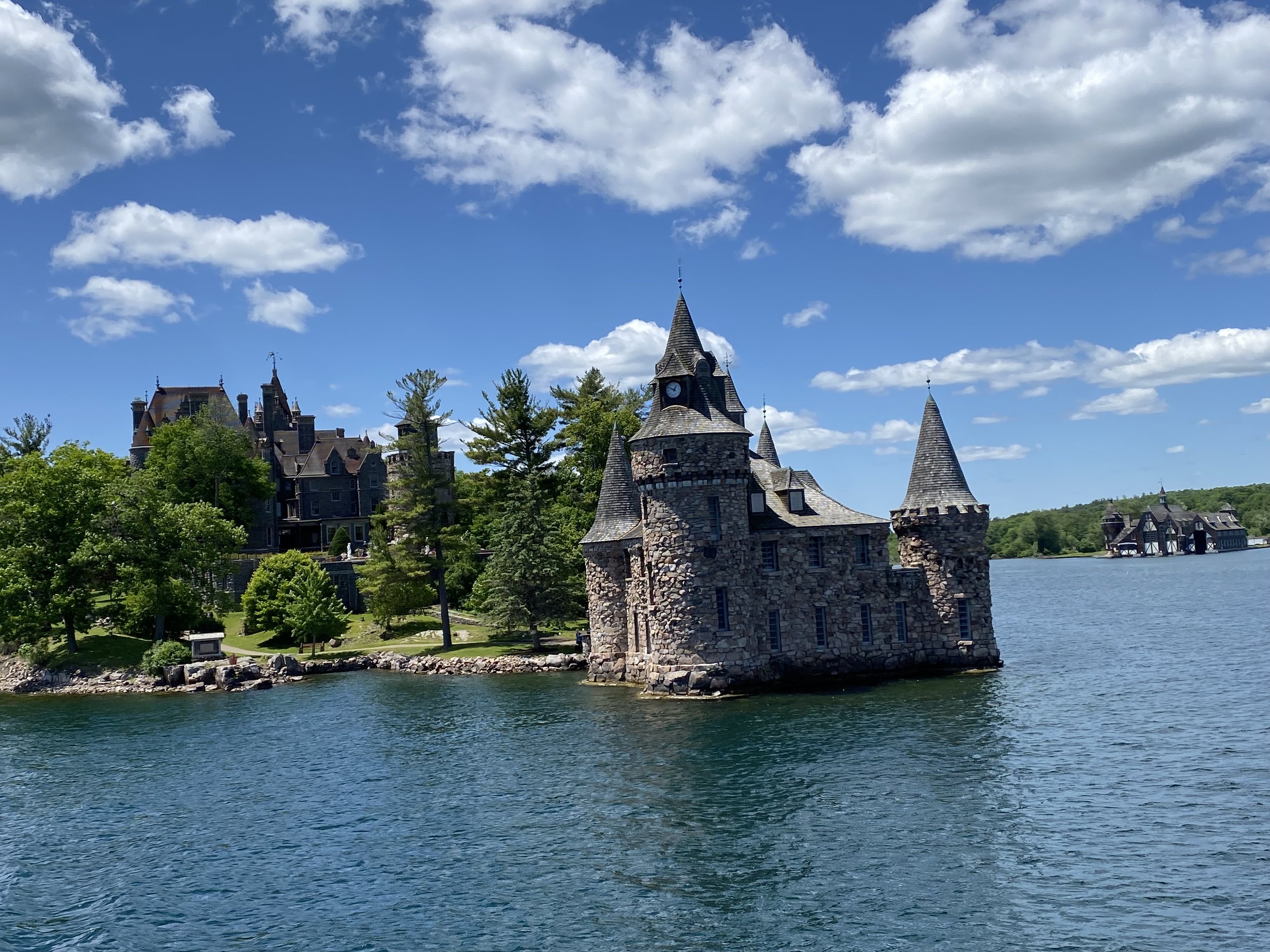 A castle with stone walls and pointed towers situated on a small island in a body of water, with green trees and a bright blue sky with scattered white clouds in the background.