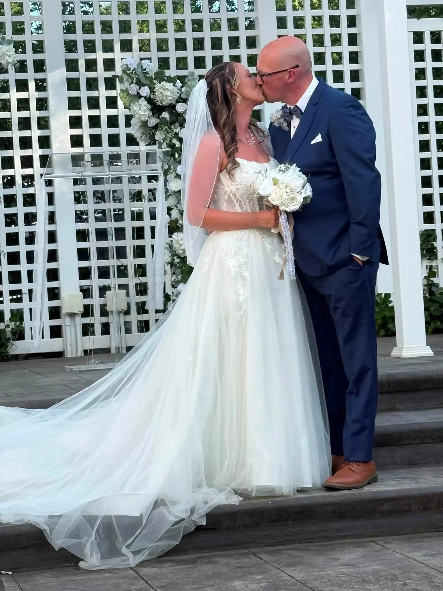 A bride and groom kiss during their wedding ceremony outdoors, with a white lattice fence and flowers in the background.