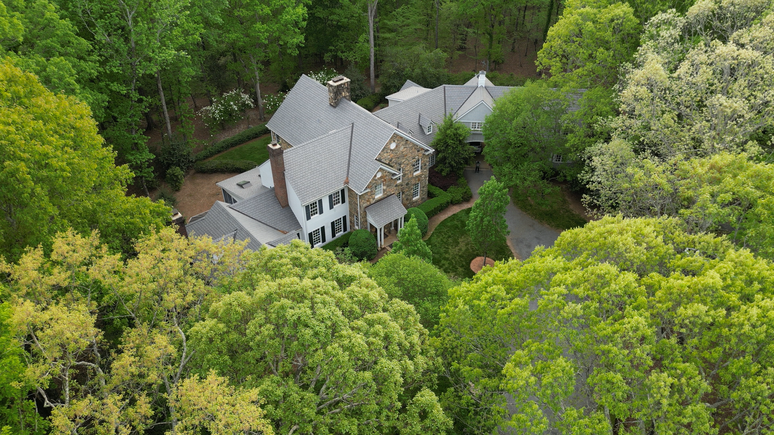 Aerial view of a large house with a gray roof surrounded by dense green trees and foliage.