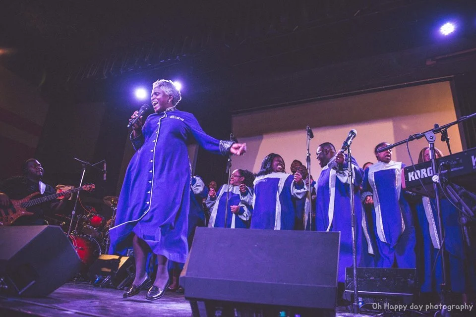 A gospel choir performing on stage directed by Karen Burke, gospel music educator, conductor and composer, in a blue robe singing into a microphone, accompanied by a guitarist and drummer, with microphones set up and stage lighting