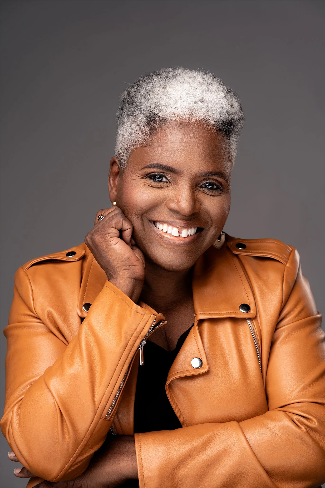 Close-up portrait of Karen Burke, Canadian gospel music educator and conductor with short, curly, gray hair, wearing a tan leather jacket, earrings, and rings, with a gray background.