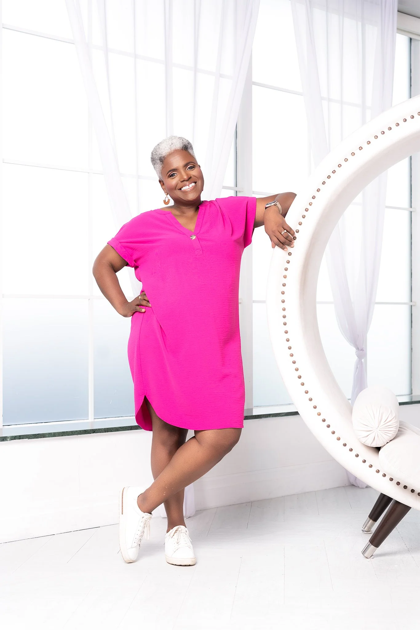 Portrait of Karen Burke, gospel music educator, conductor and composer, in a pink dress, posing with one hand on her hip and the other resting on a large white decorative object, in front of a large window with sheer white curtains.