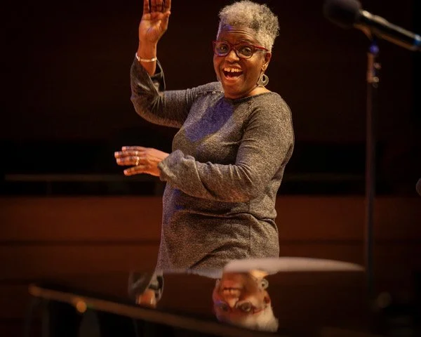 Karen Burke teaching gospel music at York University smiling and raising her hand, standing behind a piano with a reflection visible on its shiny surface.