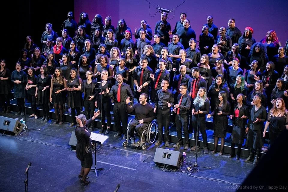 Large choir of diverse men and women singing on stage directed by conductor Karen Burke, one of Canada's leading gospel music educators. A person in a wheelchair, and stage monitors visible.
