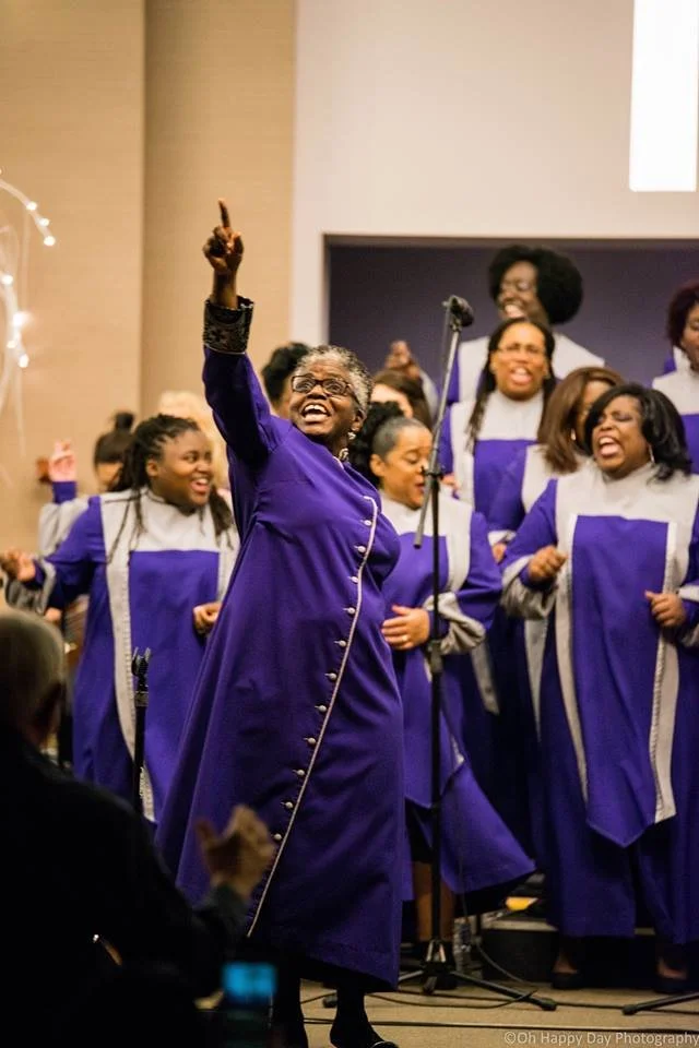 A joyful gospel choir in purple robes singing and celebrating on stage, featuring Artistic Director Karen Burke performing on stage with the Toronto Mass Choir