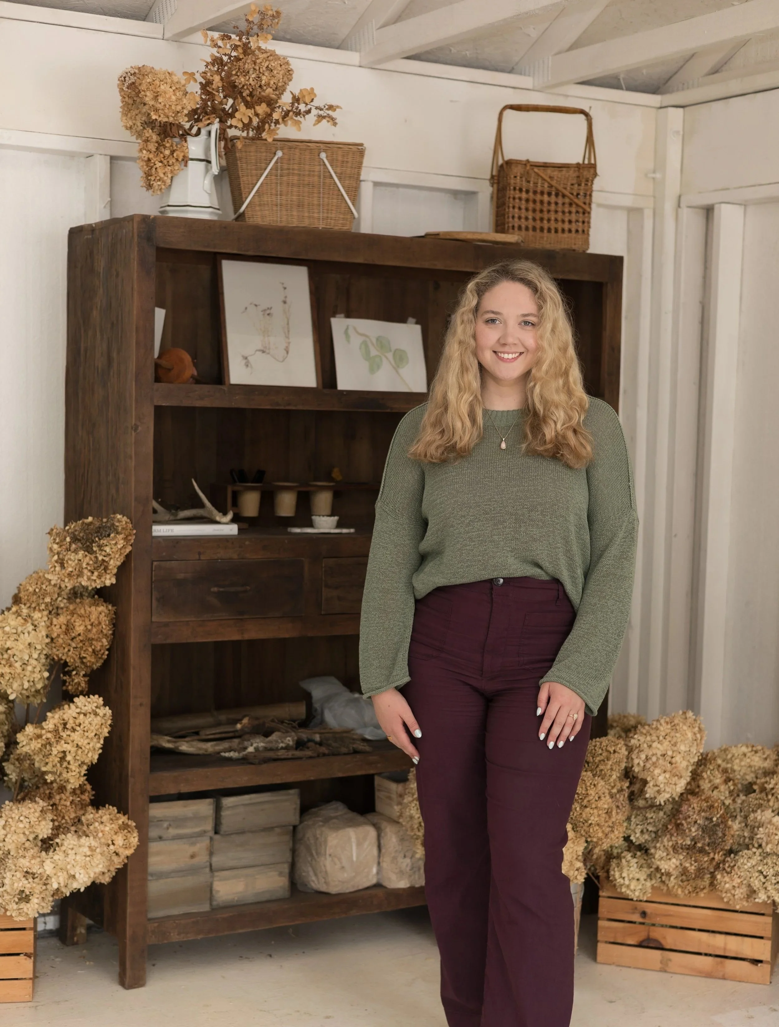 A young woman with blonde, curly hair smiling, wearing a green sweater and maroon pants, standing indoors in front of a wooden shelf decorated with framed artwork, dried flowers, and wicker baskets, with dried hydrangea flowers on the floor.