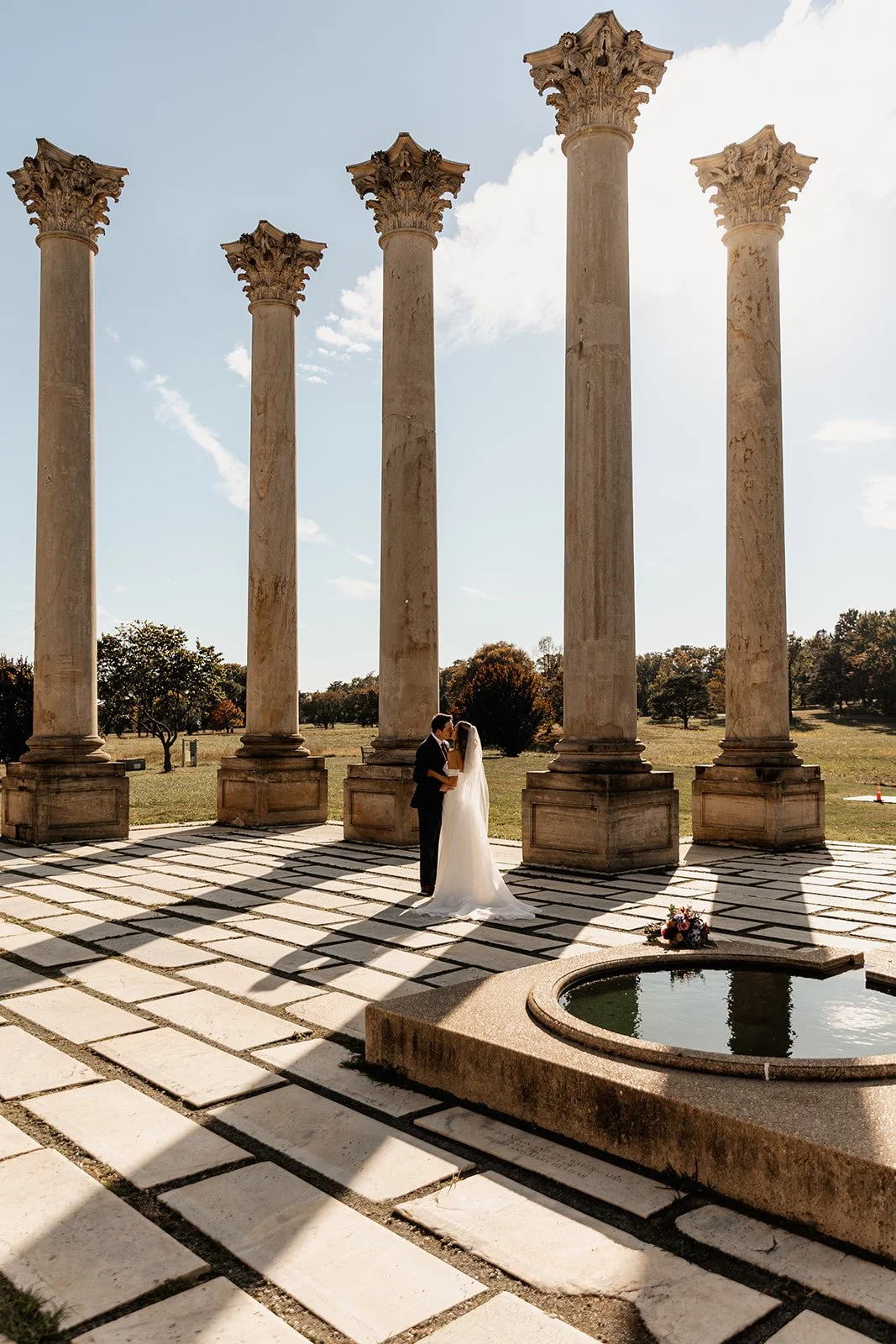 A bride and groom kissing under ancient Greek-style columns in an outdoor setting with trees and a small circular water feature in the foreground.
