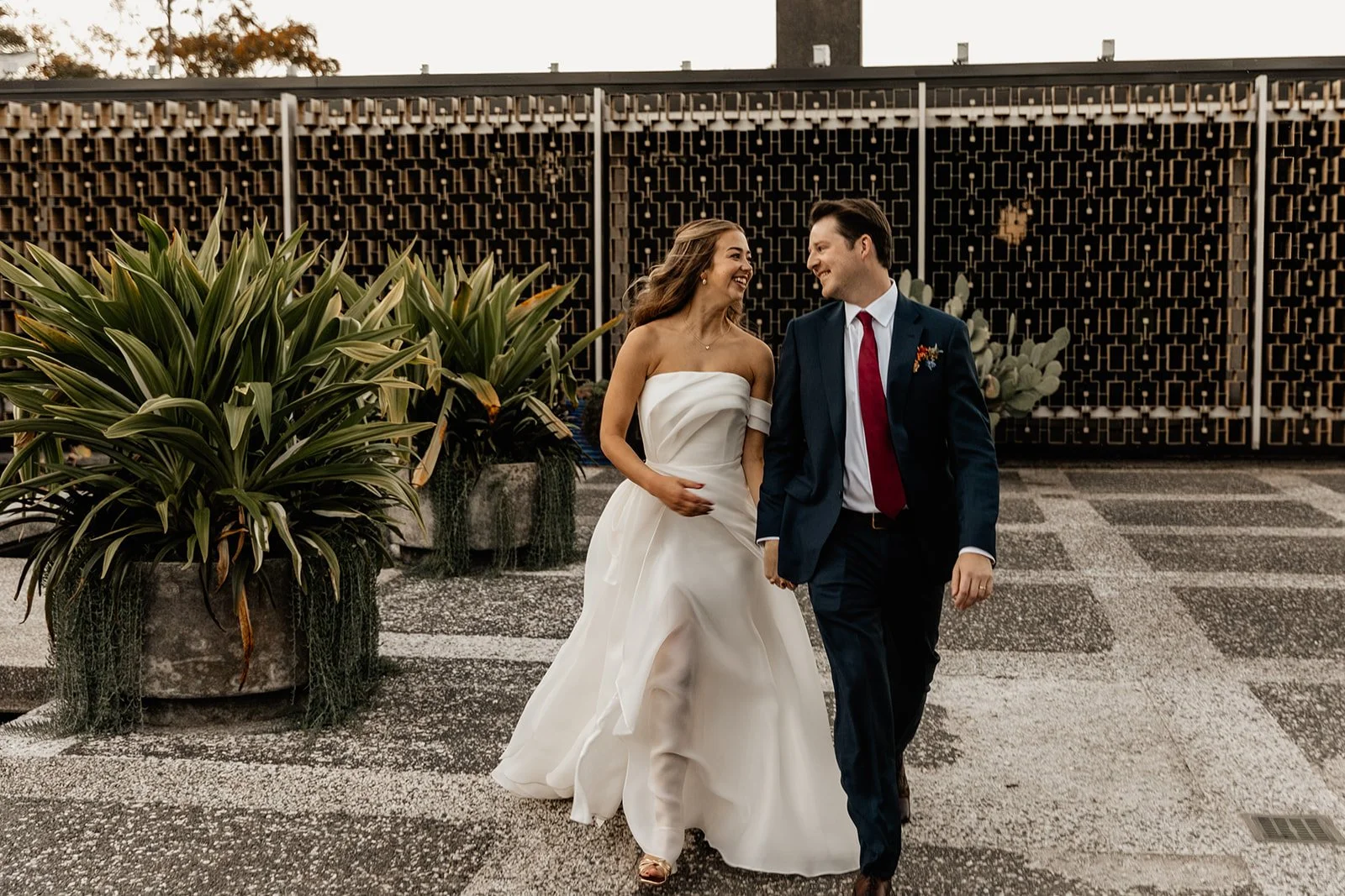A bride and groom walking outdoors during their wedding, smiling and holding hands, with greenery, decorative plants, and a modern fence in the background.