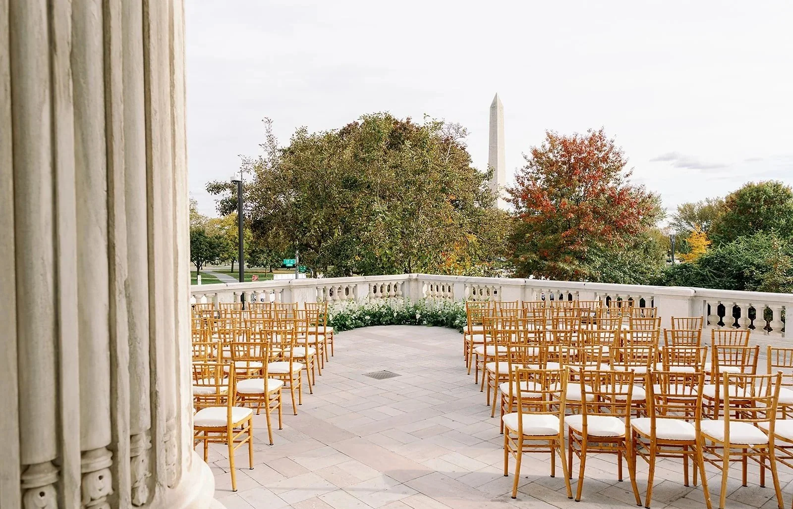 Empty outdoor balcony or terrace with wooden chairs arranged in rows, overlooking trees with autumn foliage and the Washington Monument in the distance.