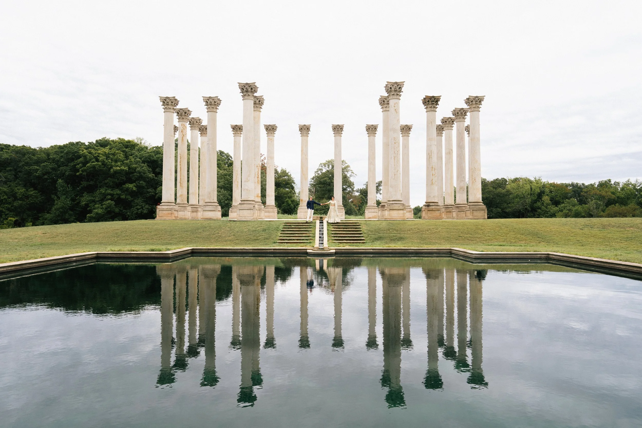 Two people standing with arms extended at the Ionian Palace ruins, ancient Greek columns, reflected in water in the foreground, green trees and cloudy sky in the background.