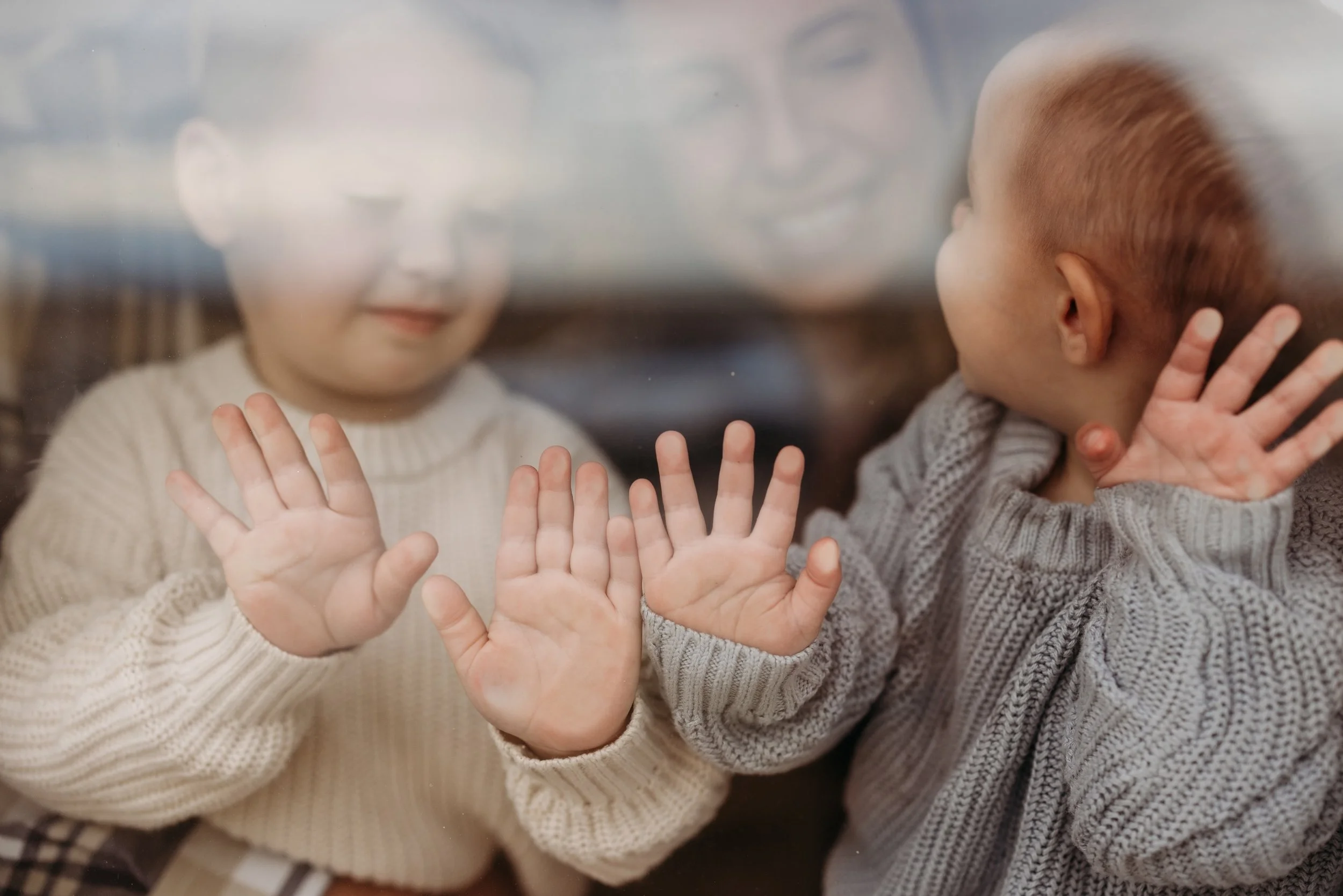 Two kids pressing their hands against a window with their mother smiling behind them.
