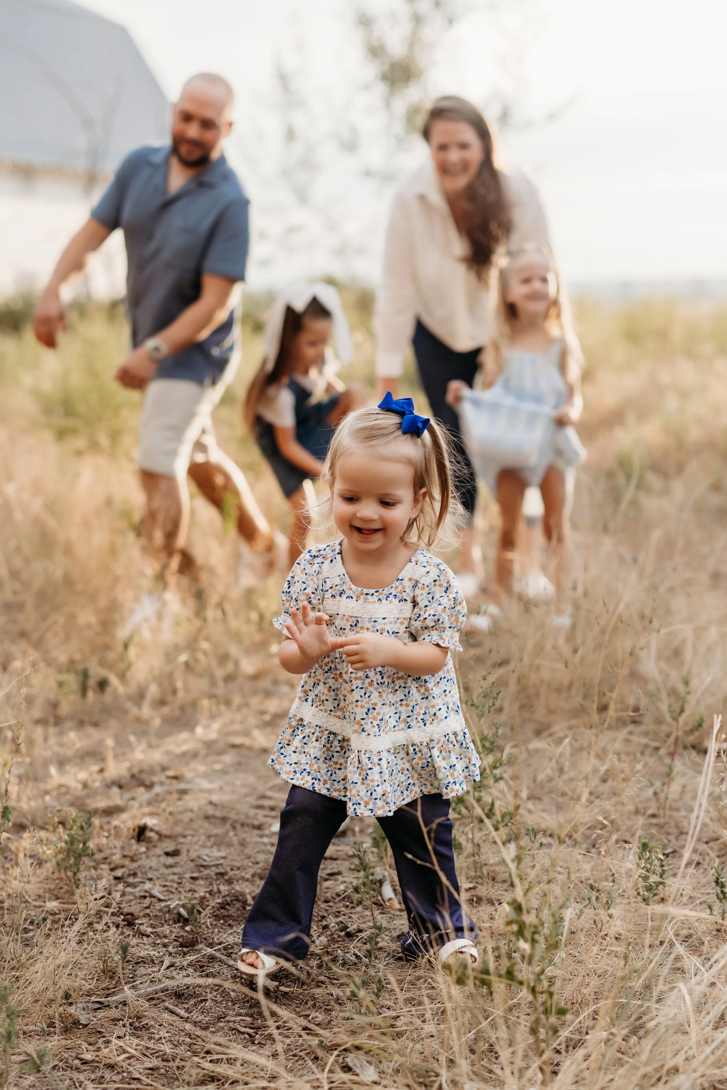 Toddler smiling with her parents and two sisters playing behind her in a field in Longmont, Colorado.