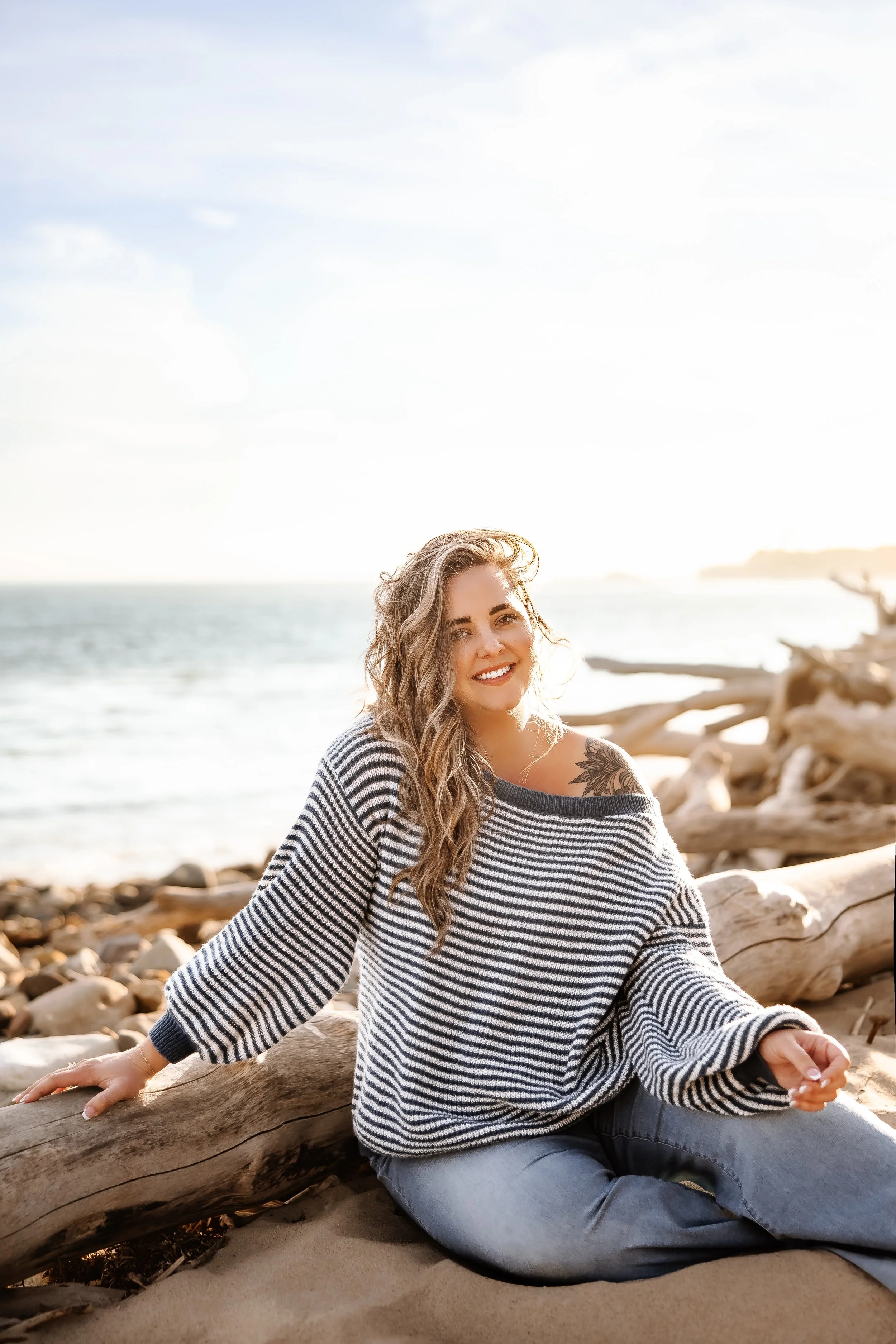 A smiling woman leaning on a piece of driftwood on a beach.