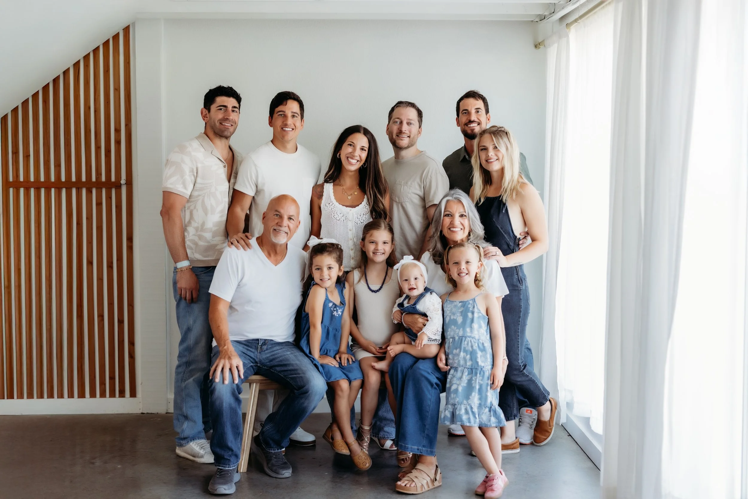 Family portrait of multiple generations, including children, parents, and grandparents, smiling indoors with natural light from windows.