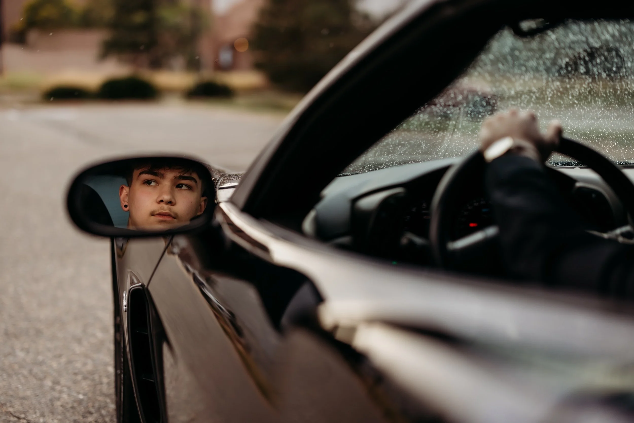 Reflection of a teenage boy in the side mirror of a black car.