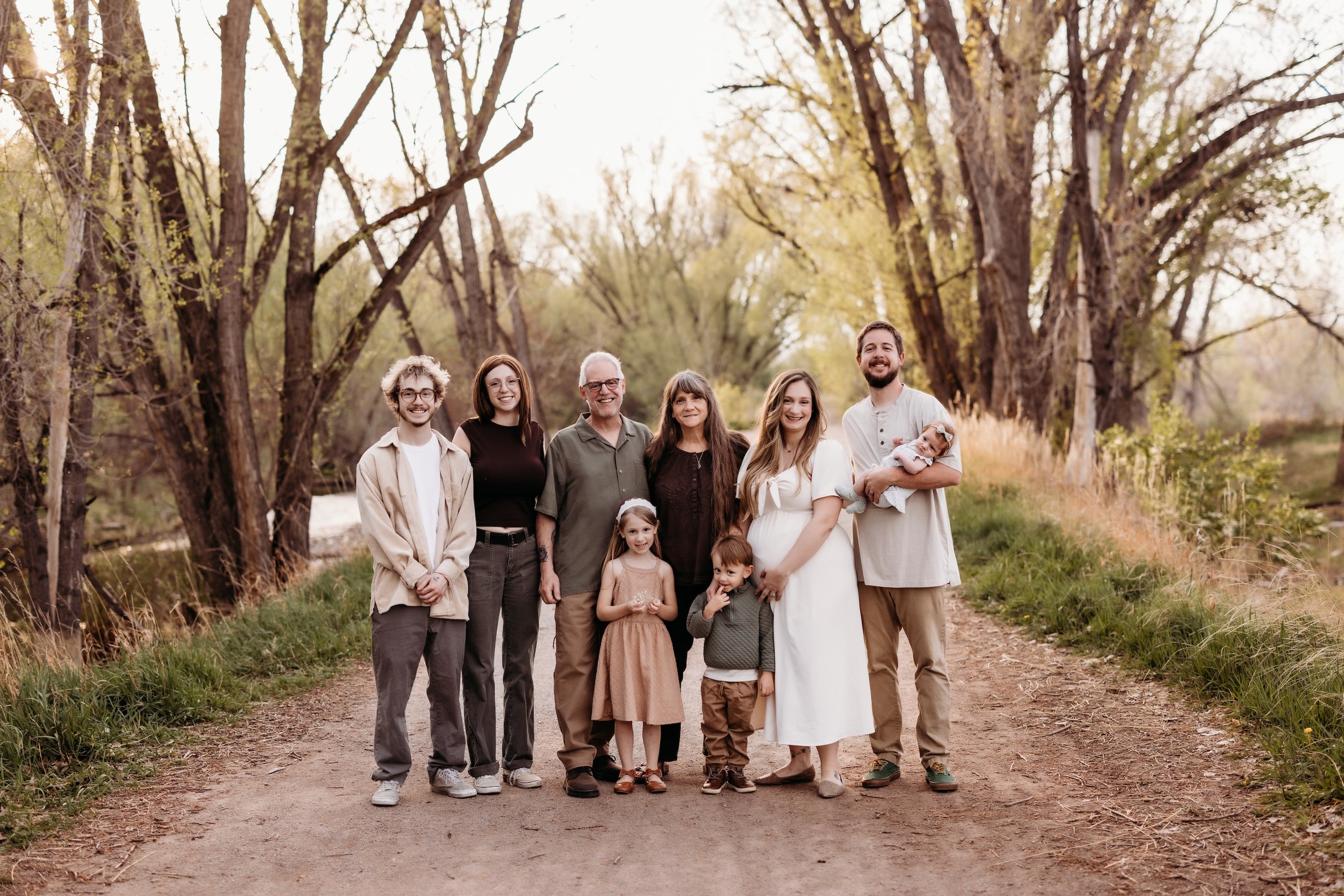 Happy family of ten, including children and adults, posing on a dirt path in a wooded area with tall trees and yellowing leaves, during autumn.