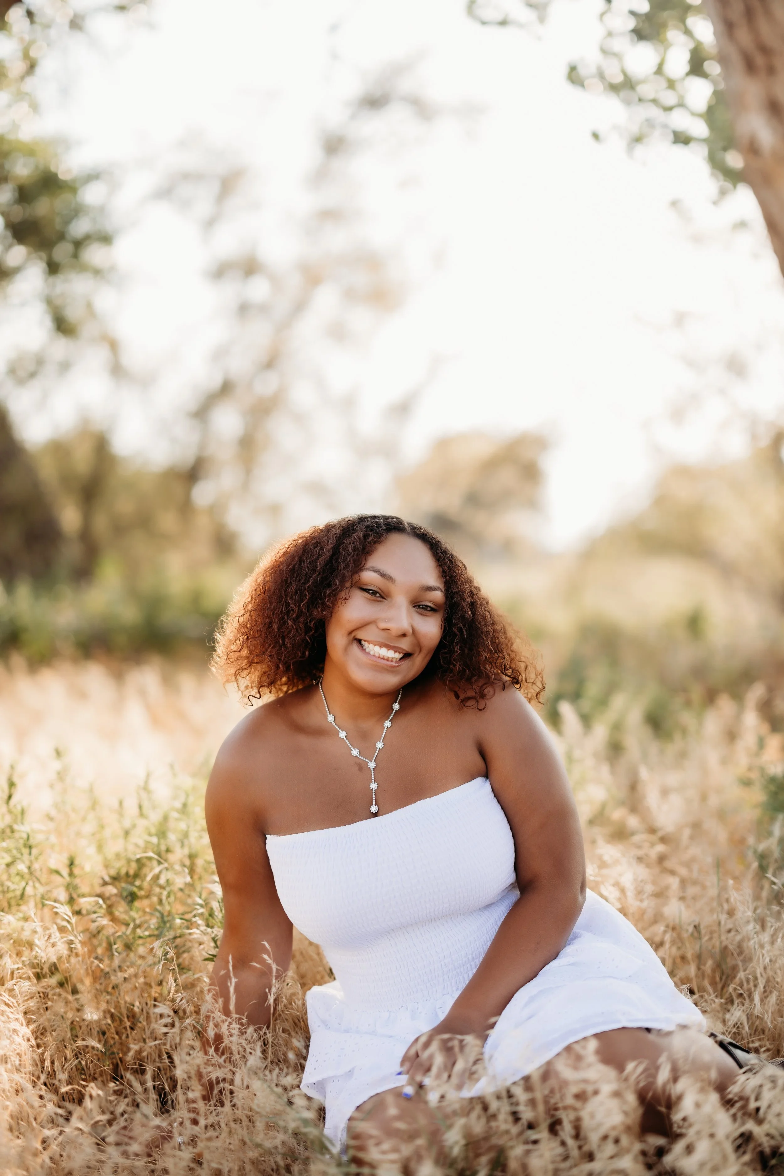 Teenage girl in a white dress smiling and sitting in a field.