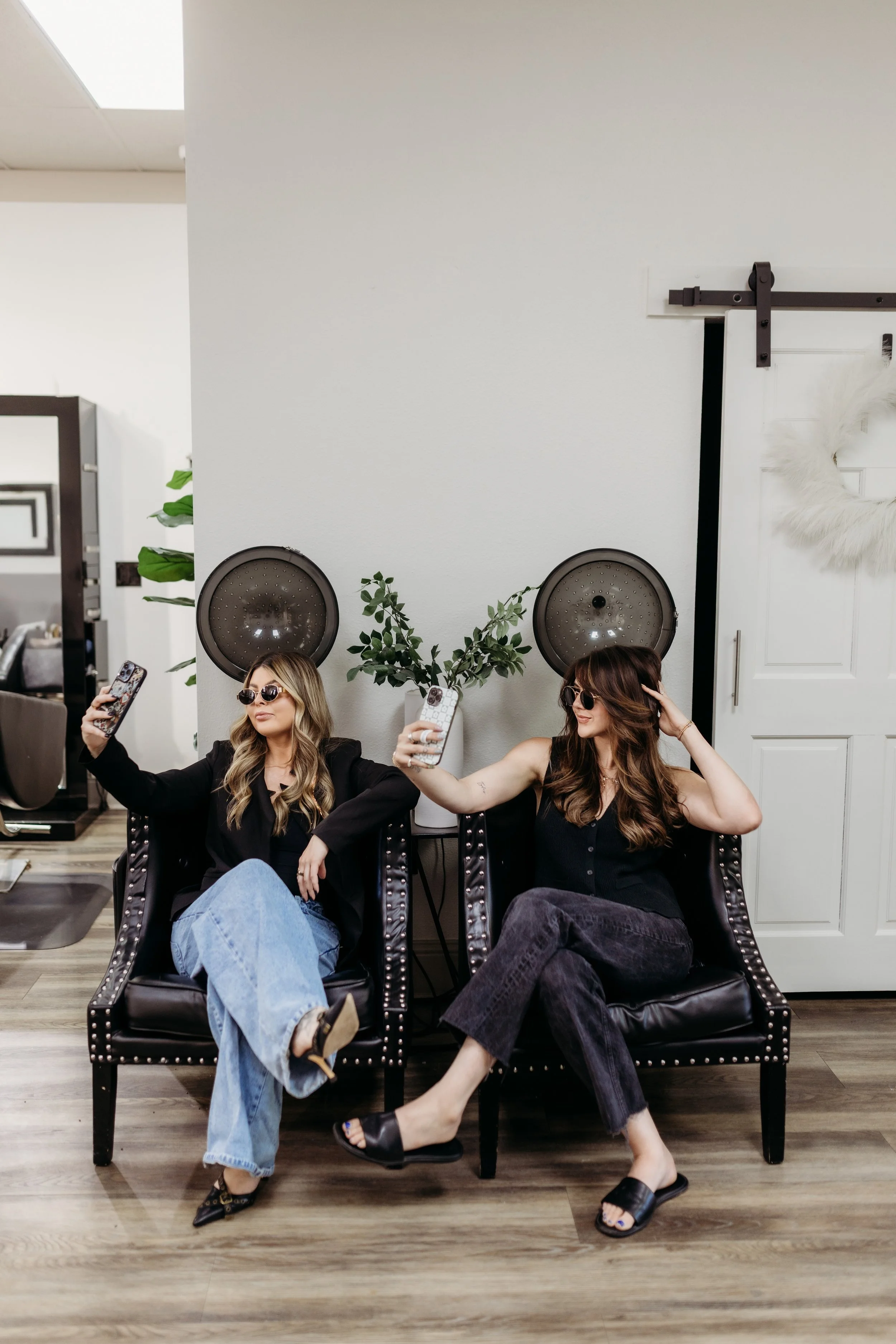 Two women taking selfies while sitting under hair dryers.