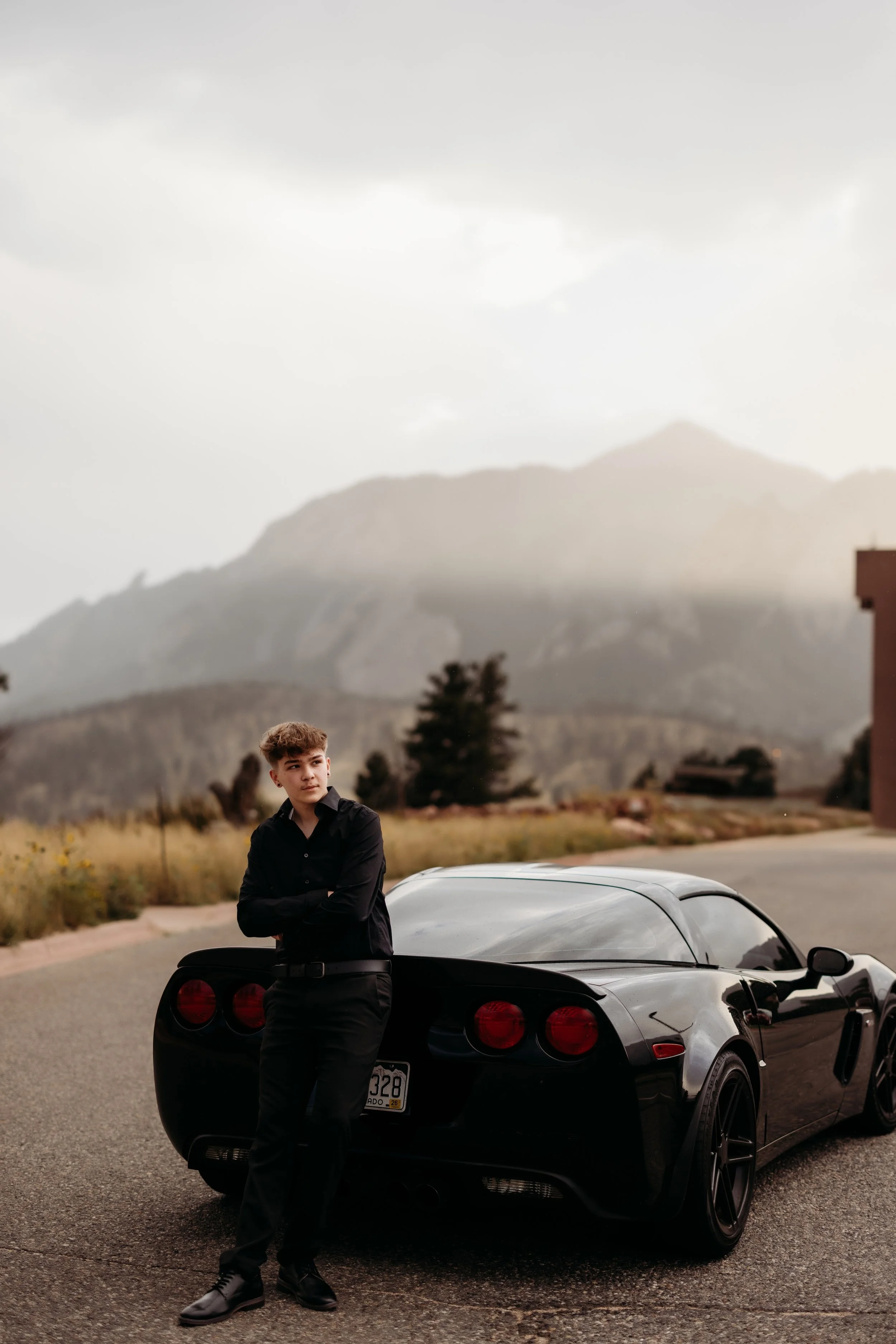 Teenage boy leaning on his car on the side of a road with a mountain range in the background.