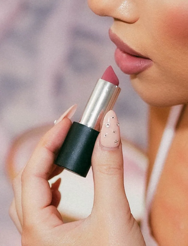 Close-up of a woman applying light pink lipstick, holding the tube near her lips, with her hand showing decorated nails with small rhinestones.