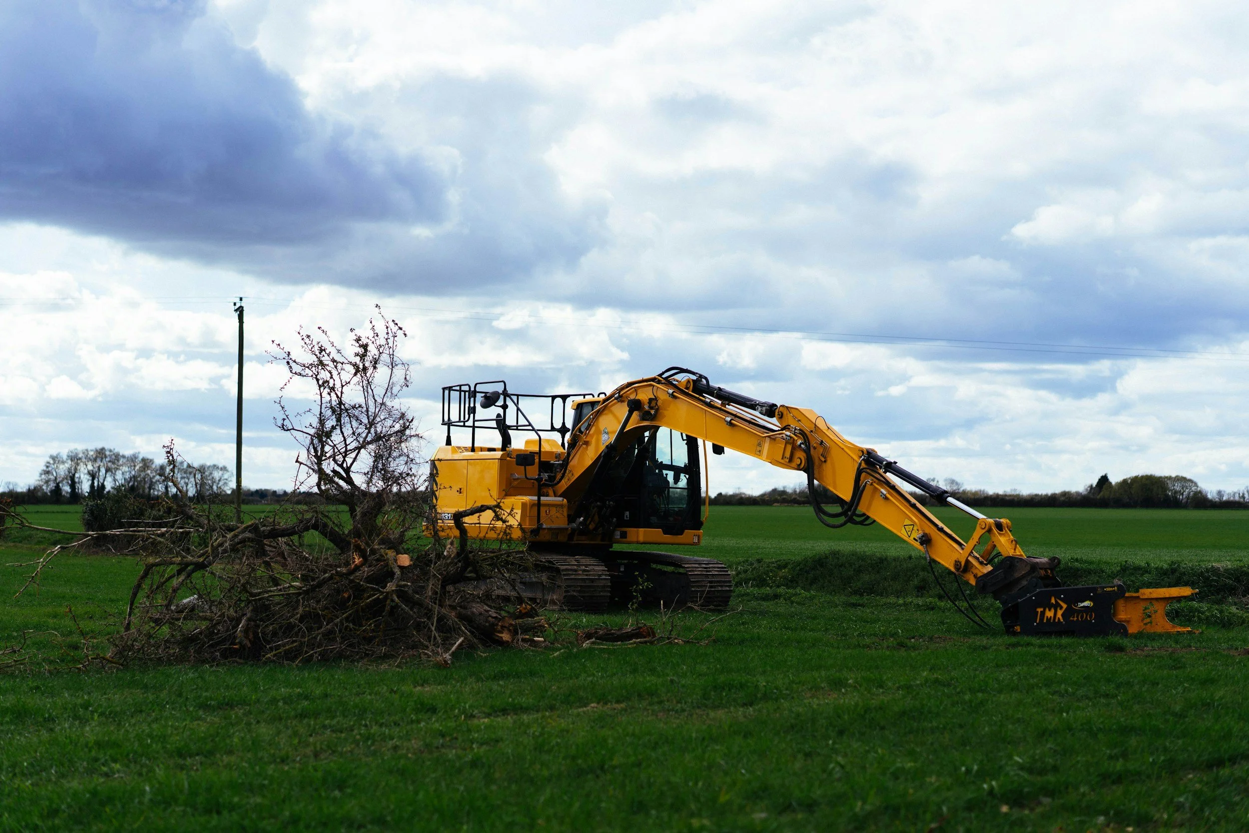 A yellow excavator moving fallen tree branches in a green field under a cloudy sky.