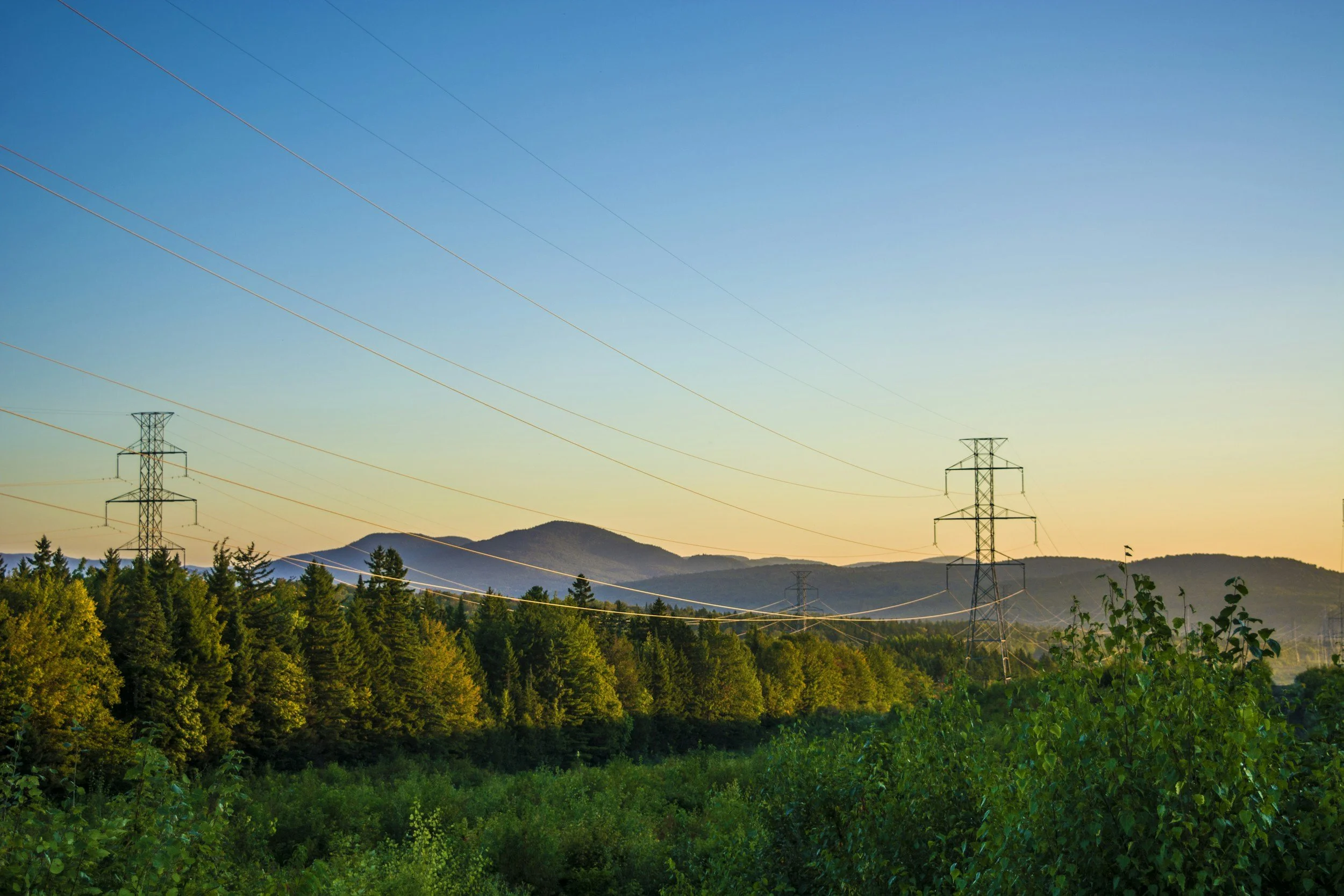 View of a landscape with green trees in the foreground, rolling hills and mountains in the background, and a clear sky with a subtle gradient from yellow near the horizon to blue higher up, featuring power lines and electrical towers.