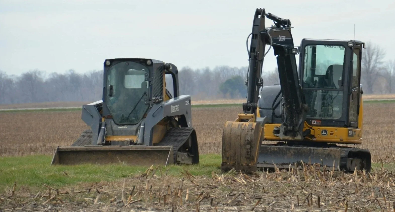 Two John Deere construction vehicles working in a field, one Compact Track Loader and one excavator, during daytime.