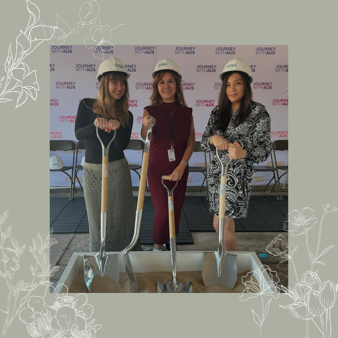 Three women wearing construction helmets and holding small shovels, posing at a groundbreaking event with a backdrop that says 'Journey With Aus.'