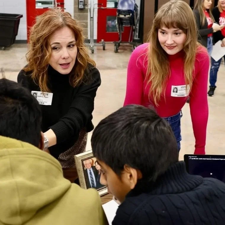 Two women with badges talking to a young man at a table with a framed photo and a laptop. Background shows other people and a red door.