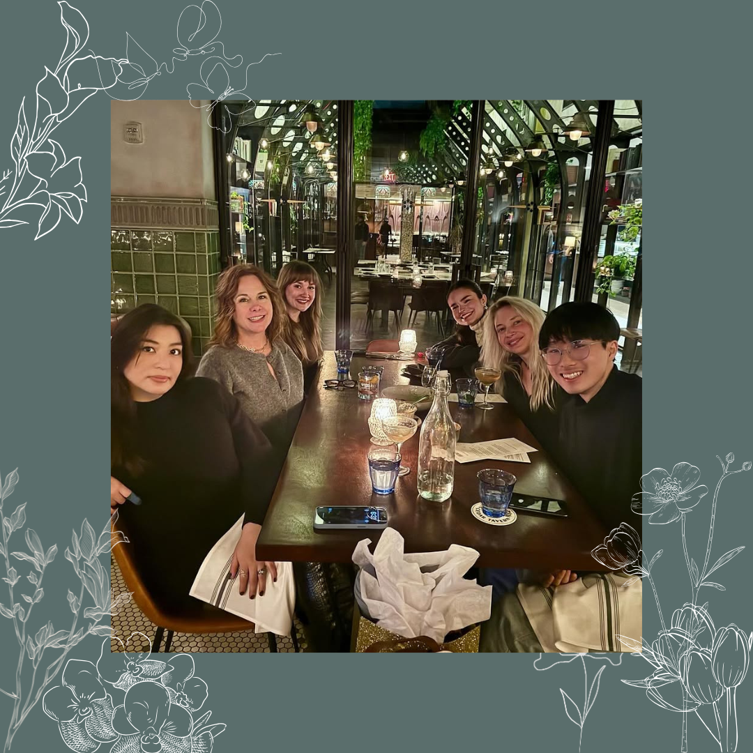 Six people sitting at a restaurant table, smiling, with drinks and a bottle in front of them, inside a well-lit dining area with glass windows and greenery outdoor view.