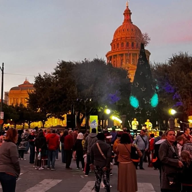Crowd gathering on a city street at dusk, with a large Christmas tree with glowing blue ornaments and a live performance stage nearby, and a domed government building in the background.