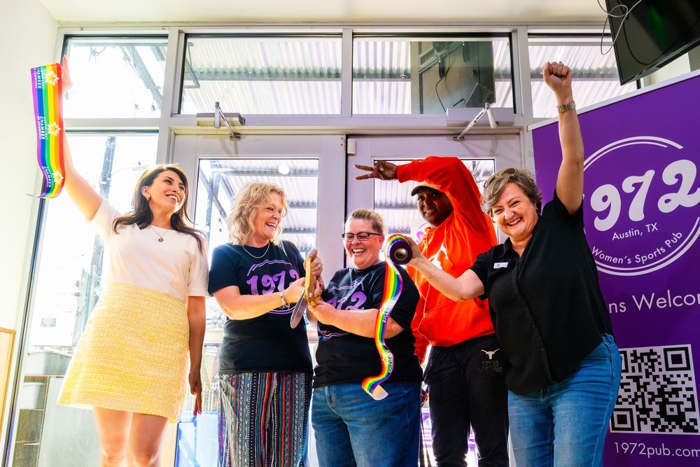 Five women celebrating inside a sports bar or pub, some holding rainbow-colored celebratory items, with a purple sign in the background that says '97.2 Austin, TX Women's Sports Pub'.