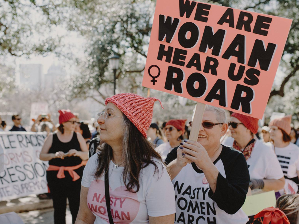Women participating in a march, holding a pink sign that reads 'We are woman hear us roar' and wearing red knit hats, with trees and other marchers in the background.