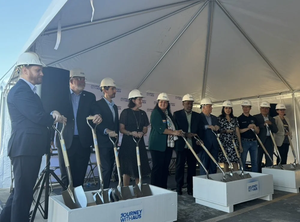People in business attire and hard hats holding shovels at a groundbreaking ceremony under a white tent.