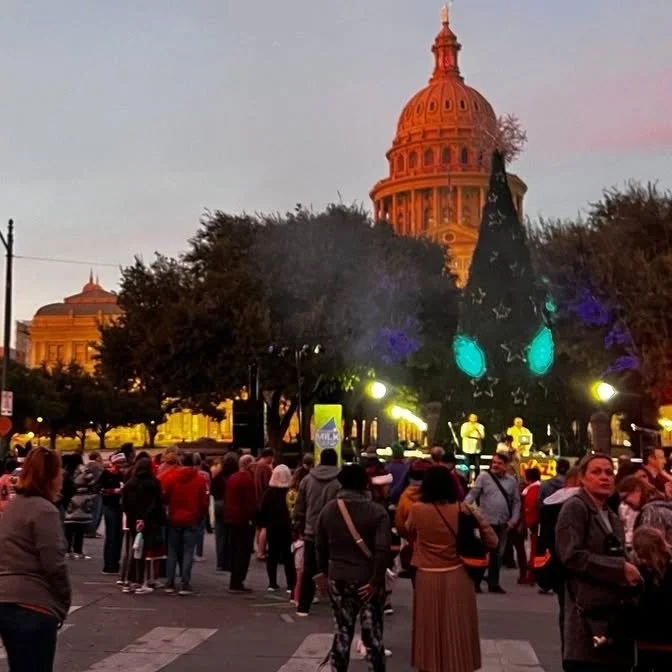 Crowd gathered around a large, illuminated Christmas tree in a city square, with the Texas State Capitol building visible in the background at sunset.