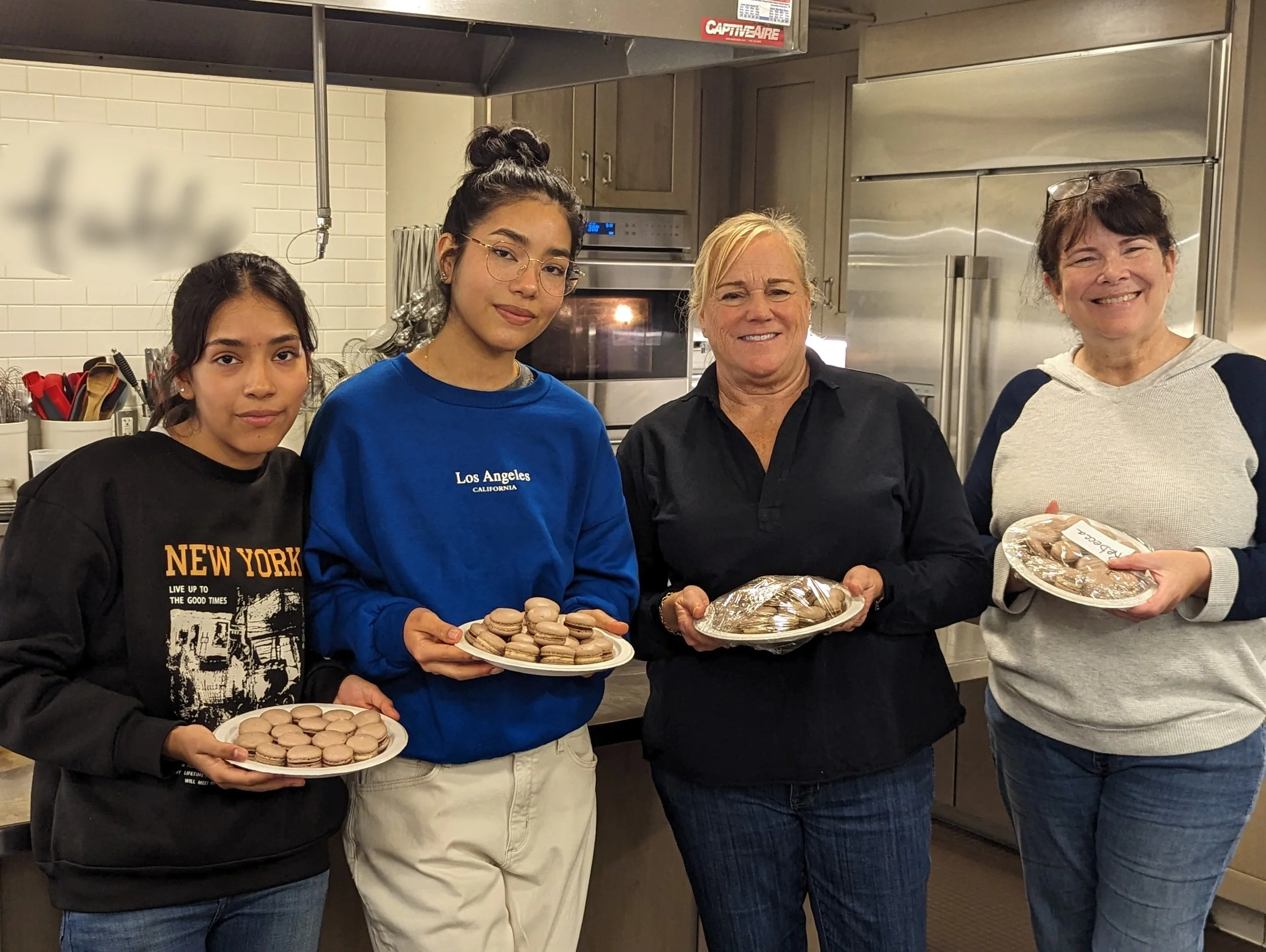 Four women, casually dressed and smiling and showing off their chocolate macarons after a successful class.