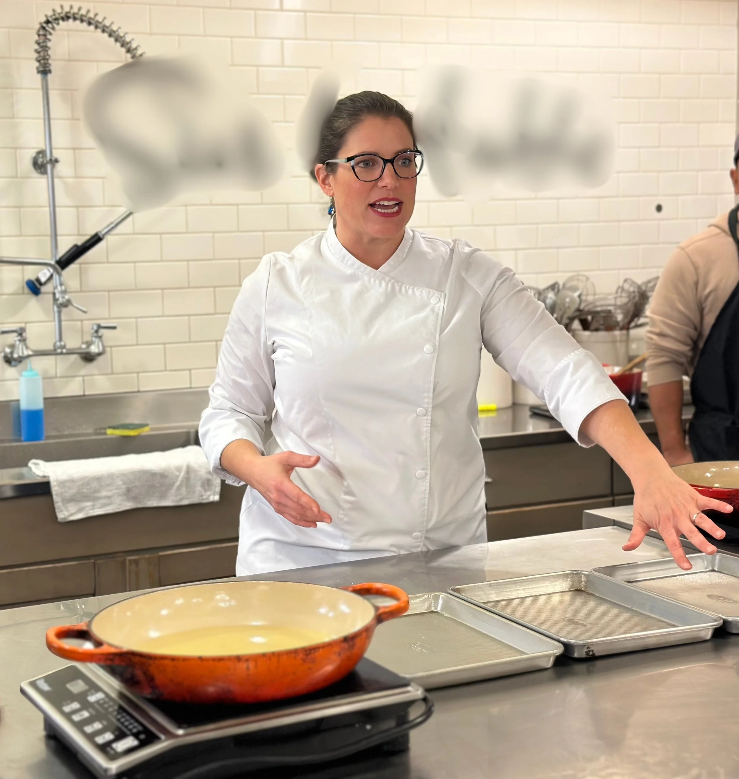 A woman in a white chef's coat and glasses is talking in a professional kitchen. In the foreground, a ceramic baking dish sits on a digital scale. The background shows a stainless steel countertop with bowls and kitchen utensils, and a brick wall.