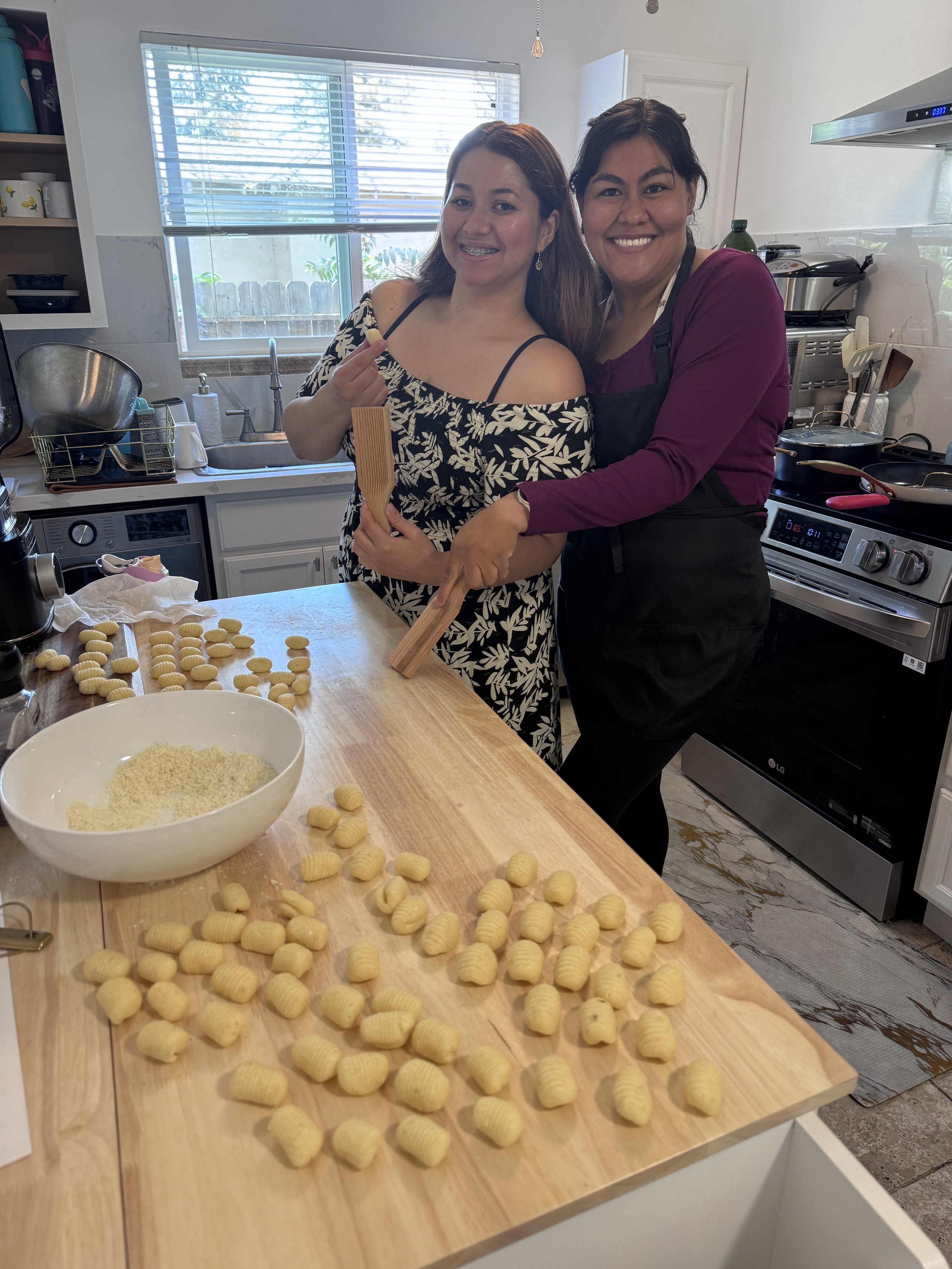 Two women smiling in a kitchen, one wearing a black and white floral dress and the other wearing a black apron and maroon top, holding wooden utensils, with homemade gnocchi on the table and a large bowl of grated cheese nearby.