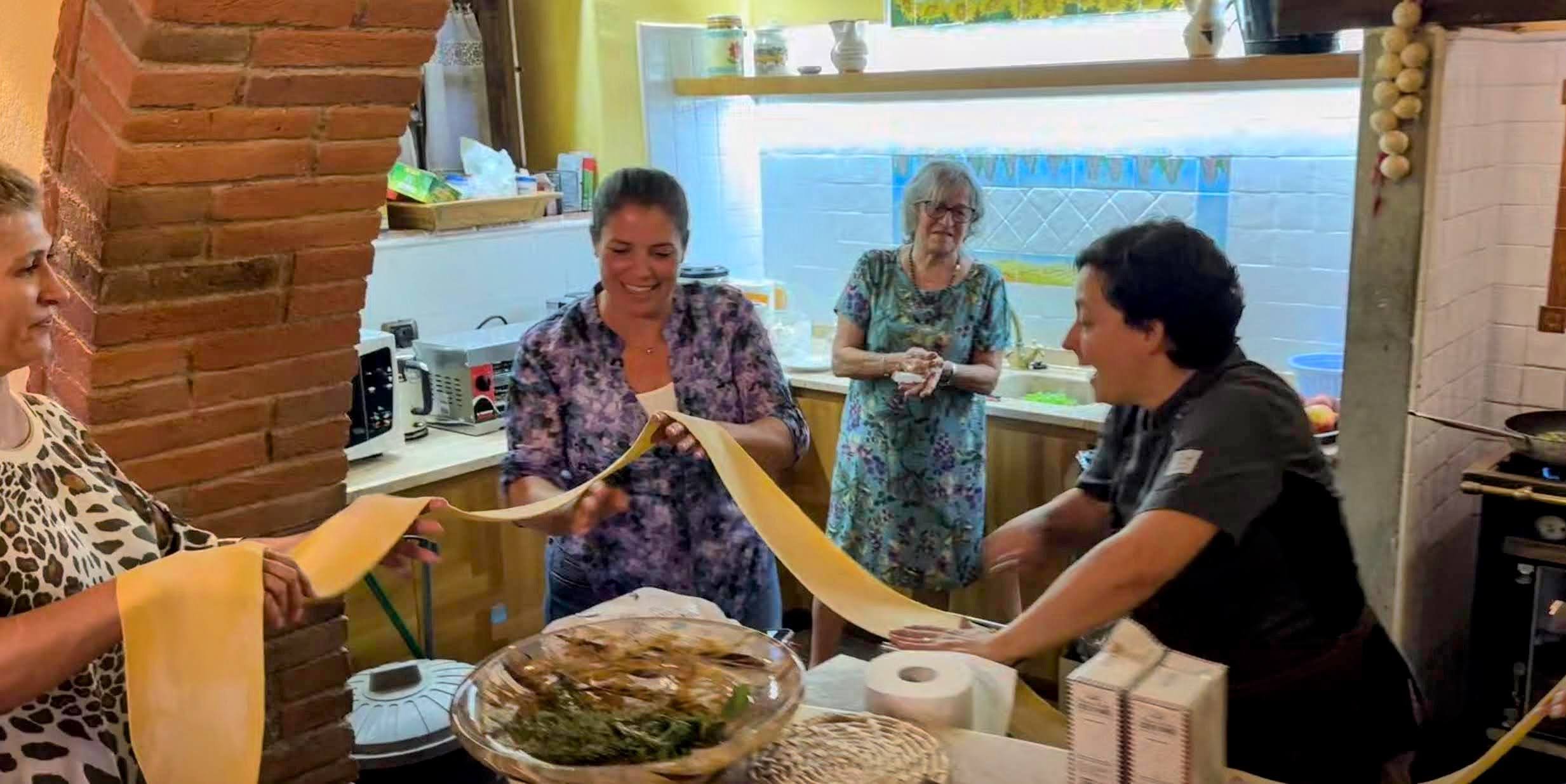Group of women and a man gathered in a kitchen preparing food, with some holding a long sheet of pasta or dough, surrounded by kitchen appliances and ingredients.