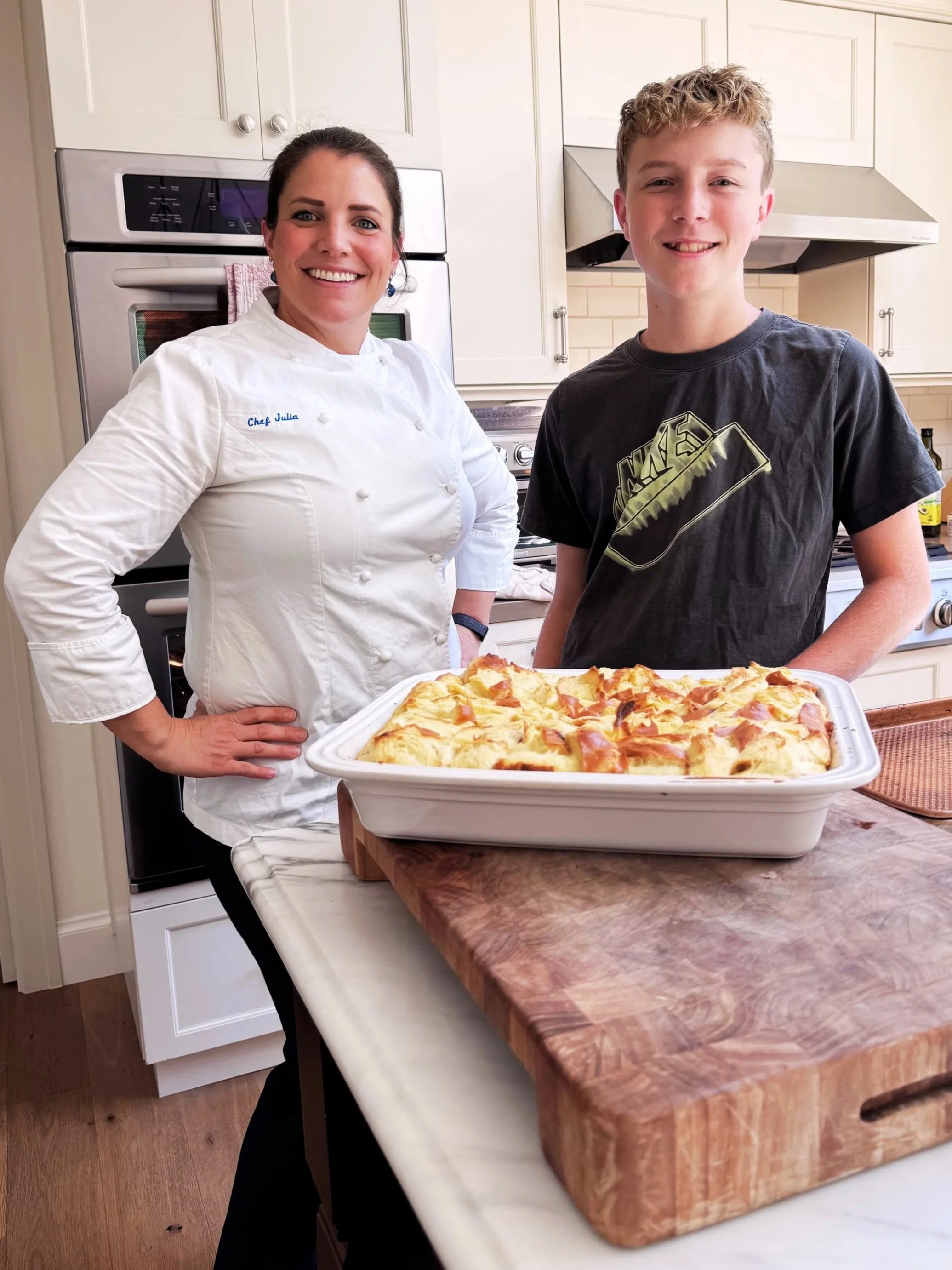 A woman in a chef's uniform and a young man stand in a kitchen, smiling. They are holding a tray of freshly baked pizza on a wooden cutting board, with a pizza oven and kitchen cabinets in the background.