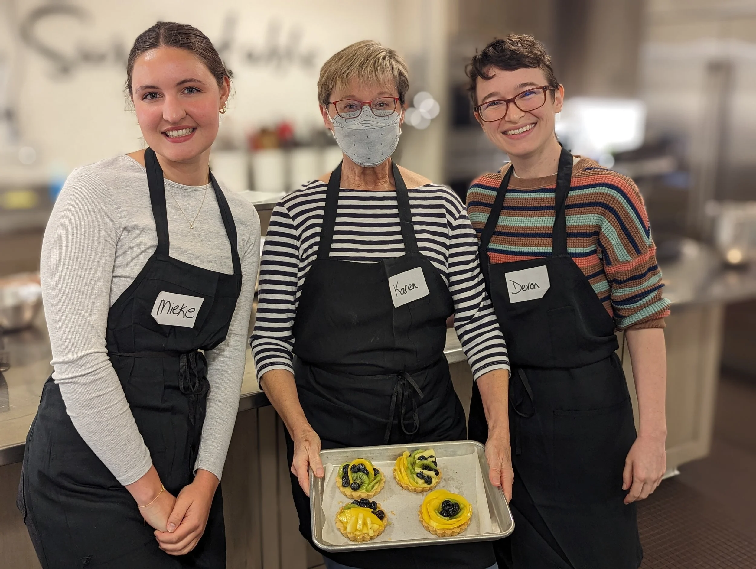 Three women wearing black aprons with name tags standing in a kitchen, one holding a tray of fruit tarts topped with kiwi, blueberries, and mango.